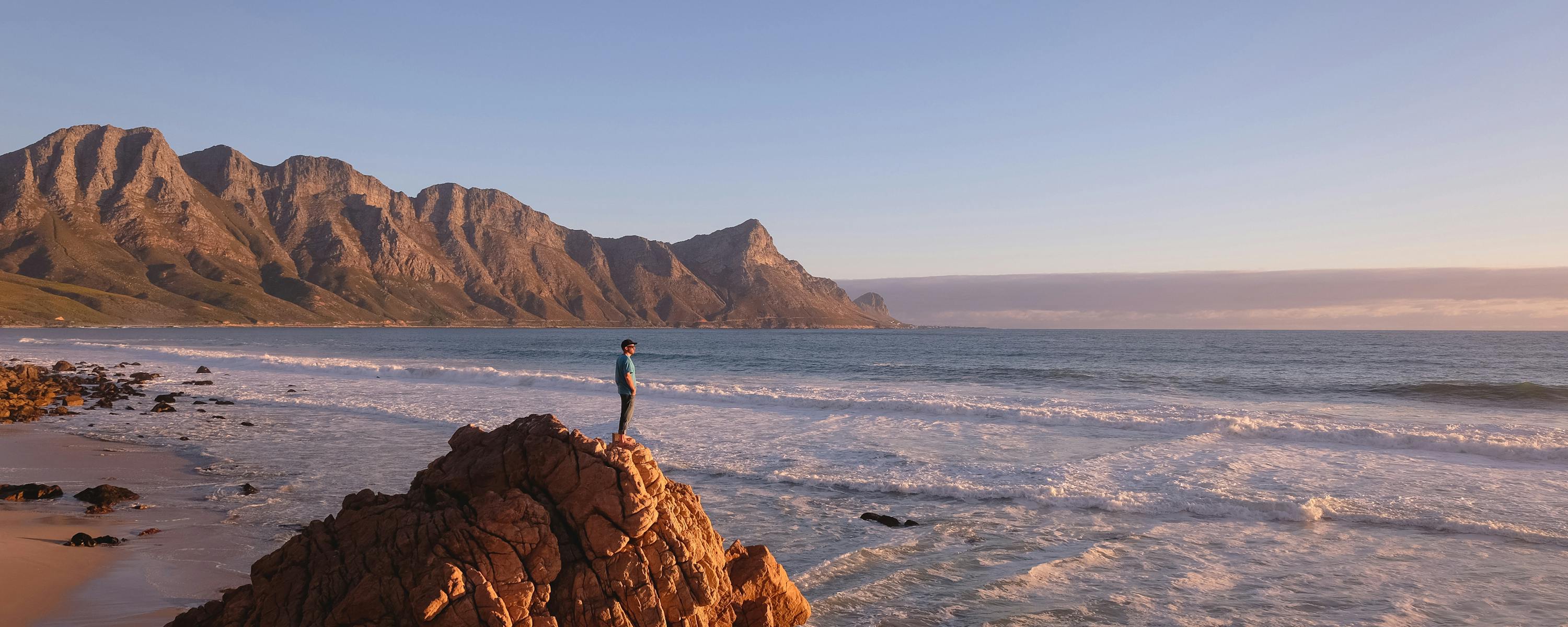 A lone traveler stands on coastal rocks above rolling surf, with a long sandy bay and mountains under pastel sky.
