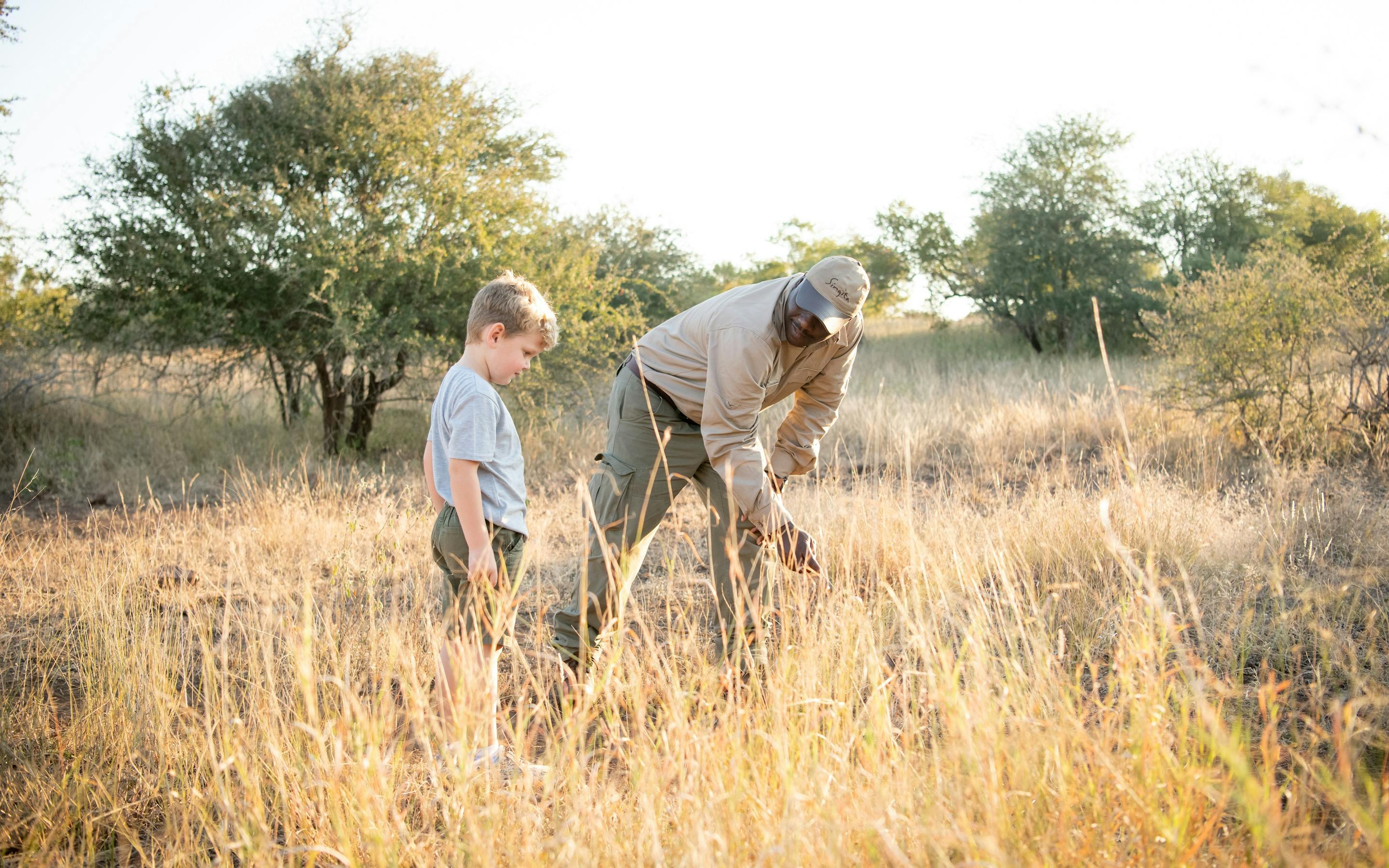 An adult guide and a child walk through tall dry grass on a bush walk, with scattered trees and golden light.