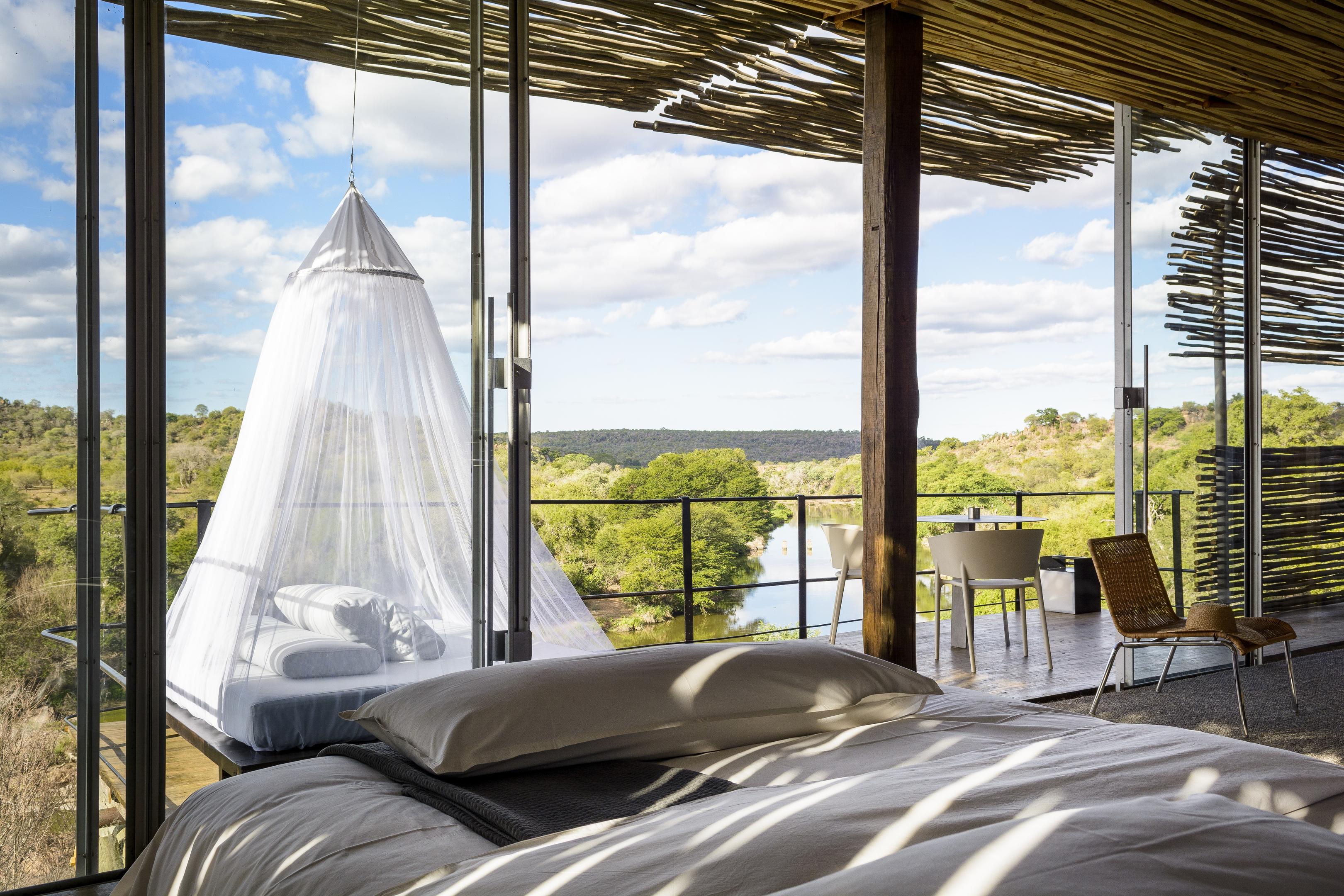 A canopy bed with netting faces an open deck at Singita Lebombo Lodge, overlooking green treetops and distant hills.