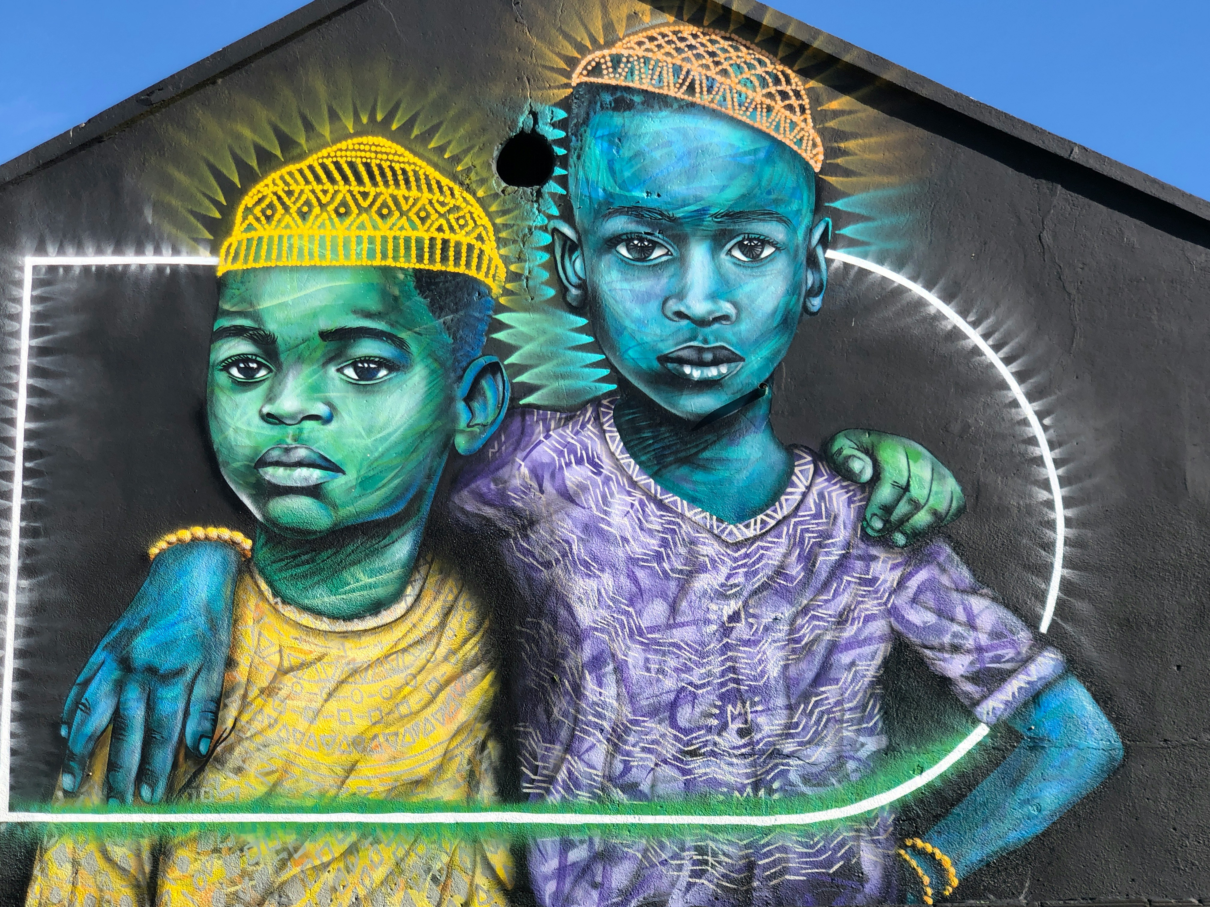 Colorful street mural shows two children with painted faces, bright halos, and patterned clothing on a wall.
