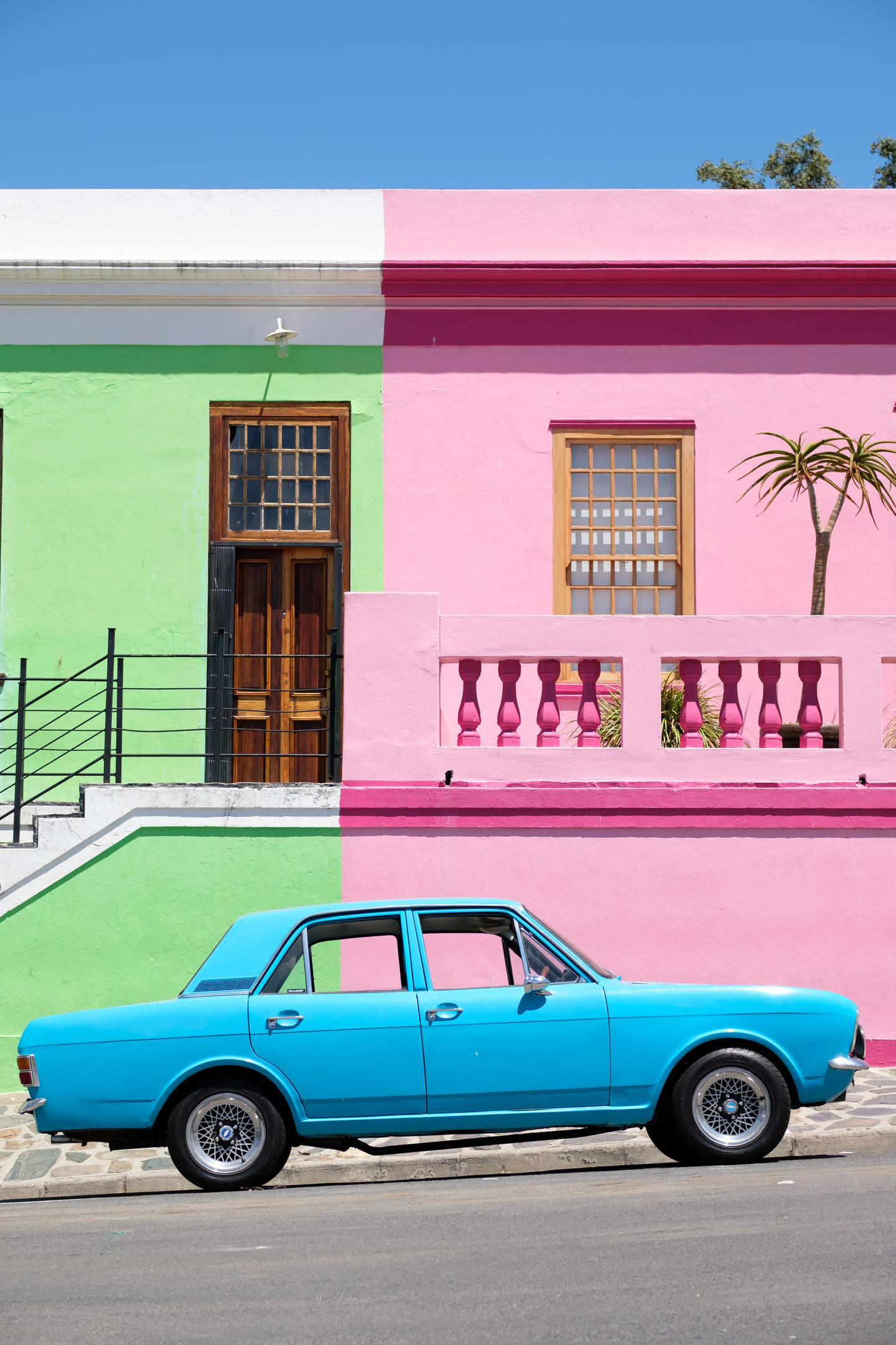 Bright Bo-Kaap houses line a steep street as a turquoise vintage car is parked beside the painted facades.