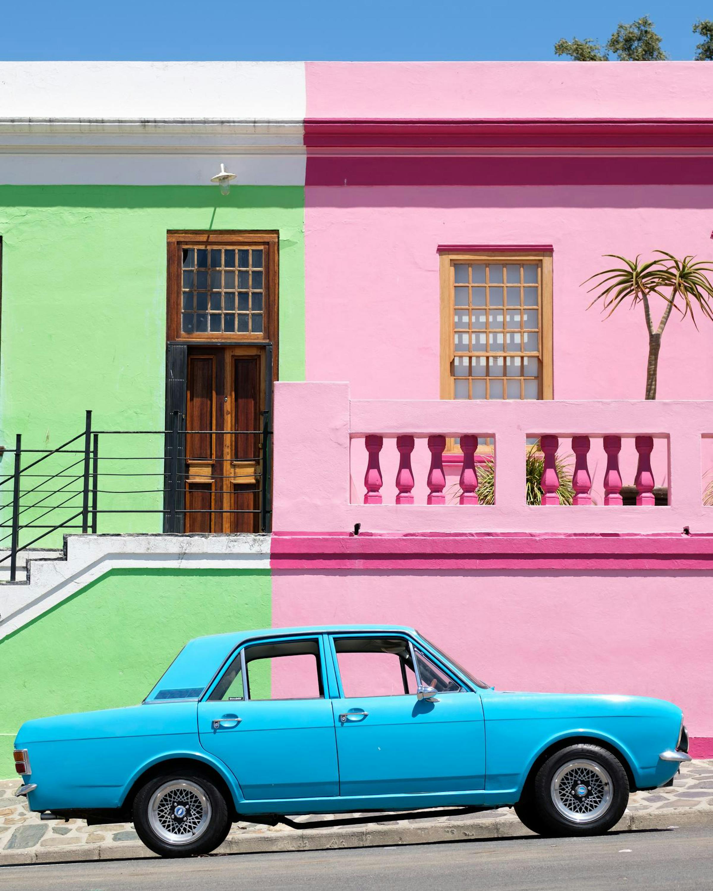 Bright Bo-Kaap houses line a steep street as a turquoise vintage car is parked beside the painted facades.