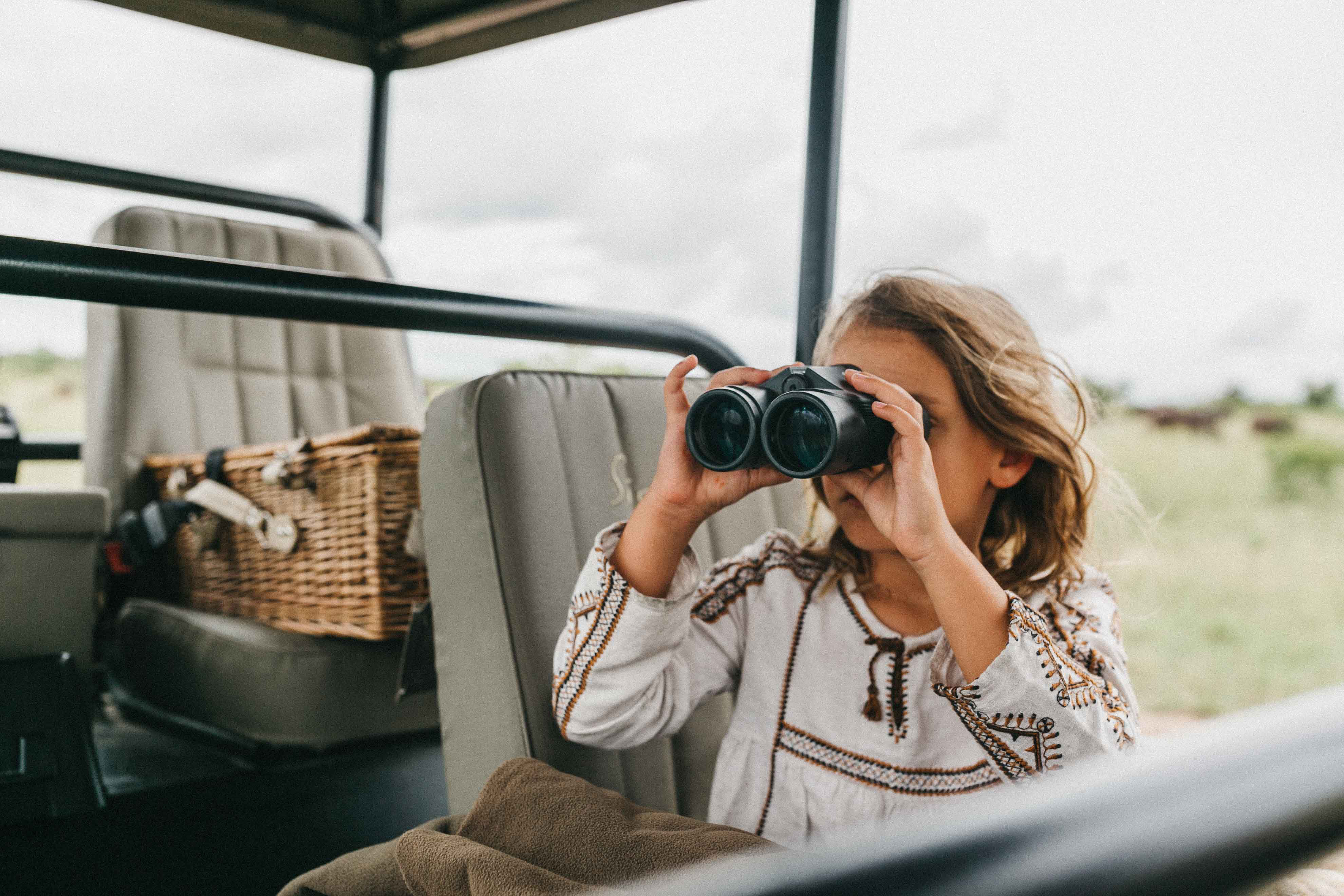 A young girl peers through binoculars from a safari vehicle, scanning the bush for animals.