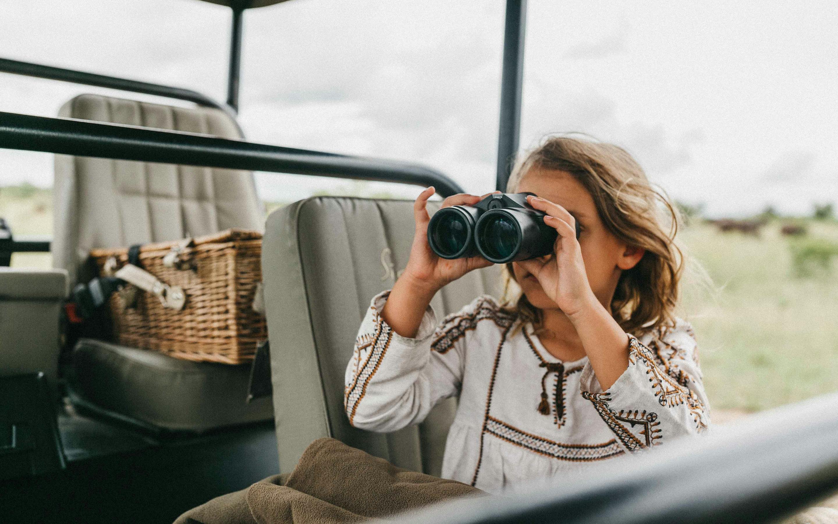 A young girl peers through binoculars from a safari vehicle, scanning the bush for animals.