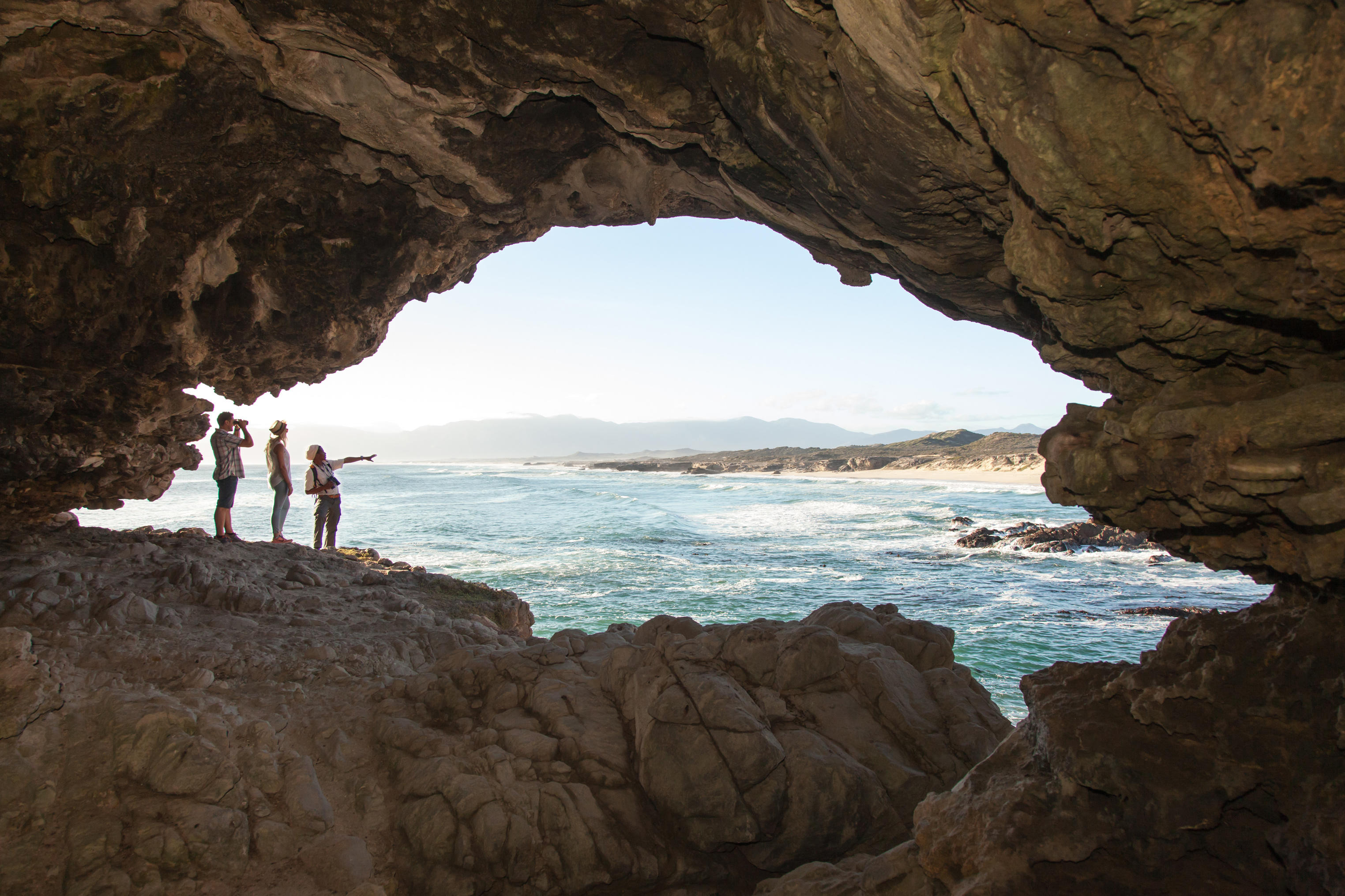 Visitors stand inside a coastal cave, silhouetted against bright ocean light and a rocky shoreline beyond.