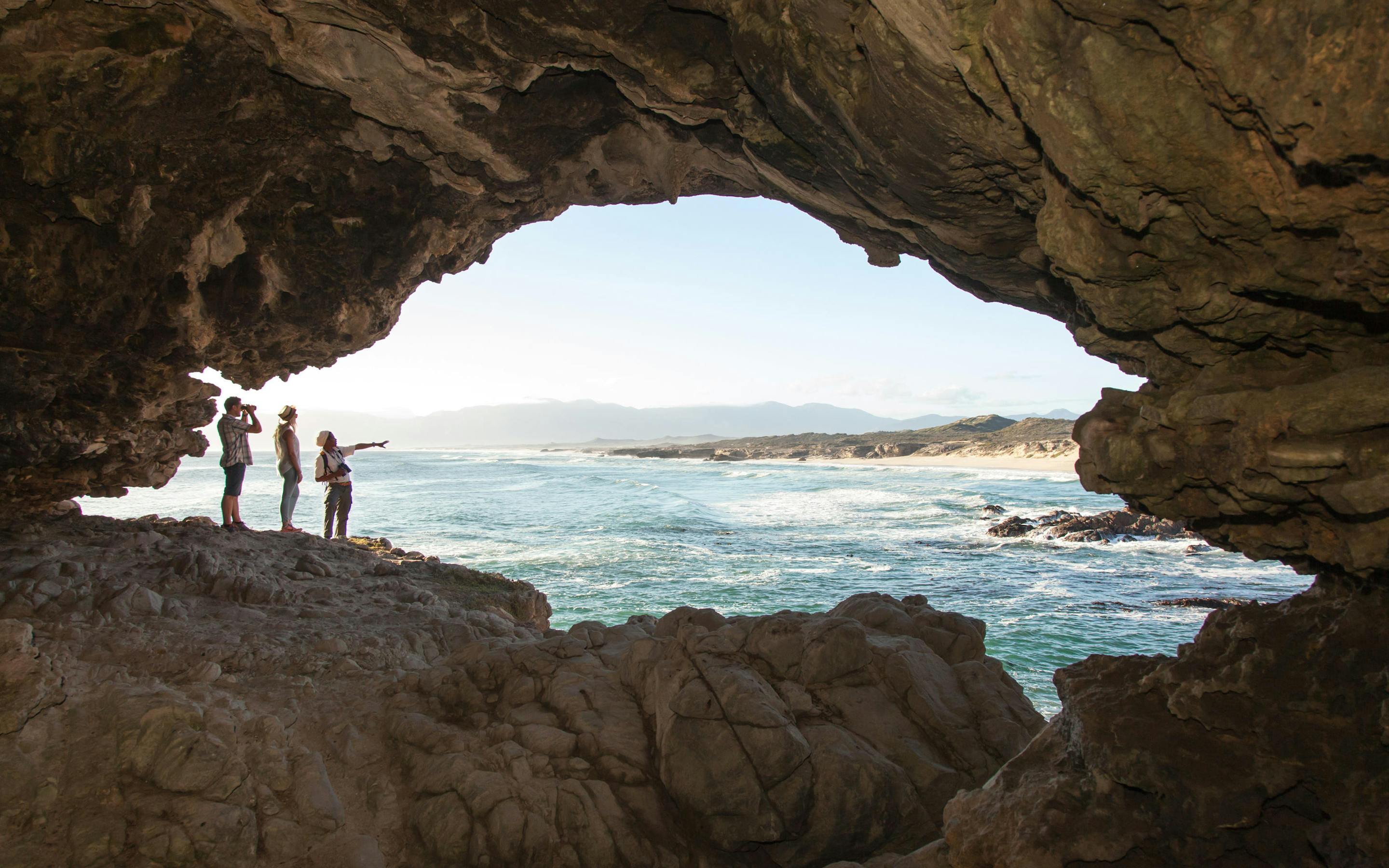 Visitors stand inside a coastal cave, silhouetted against bright ocean light and a rocky shoreline beyond.