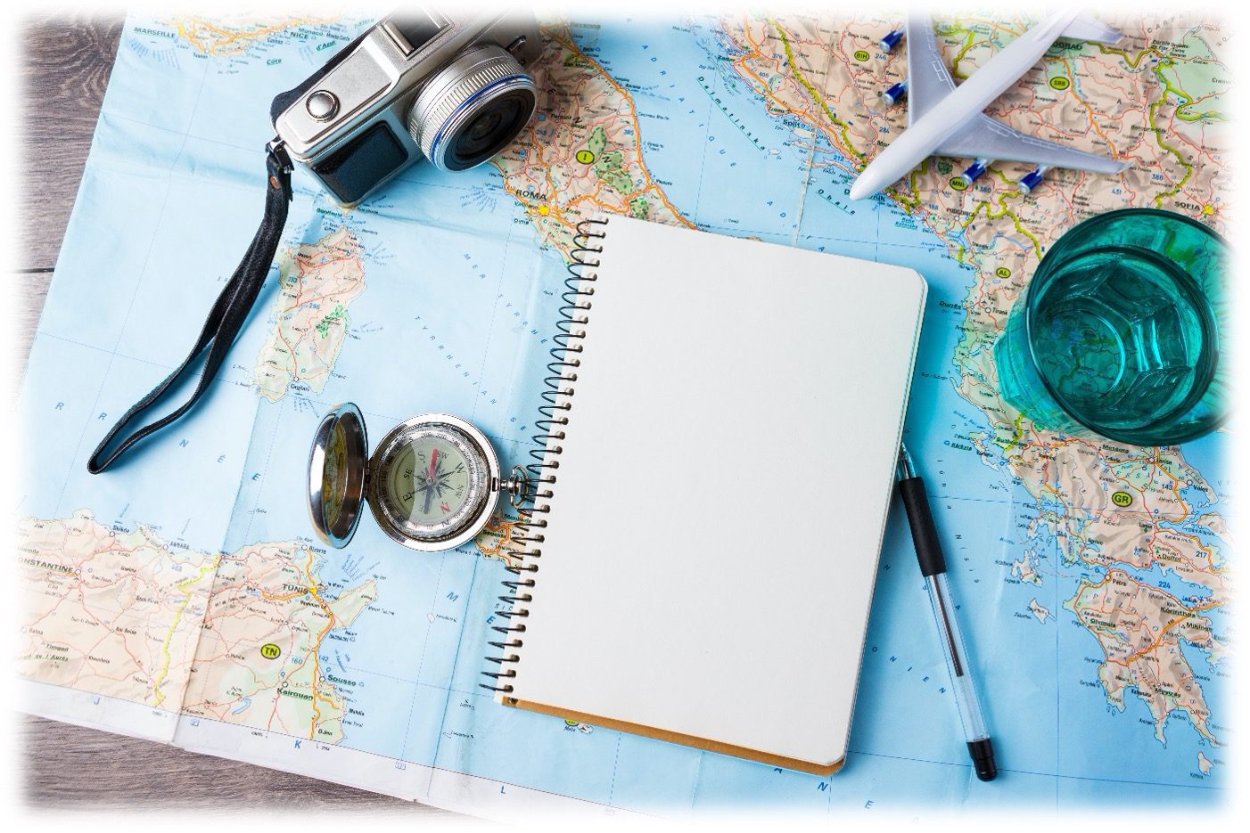 Travel flat lay with a map, notebook, passport and sunglasses, plus a camera, pen and drink on a white table.