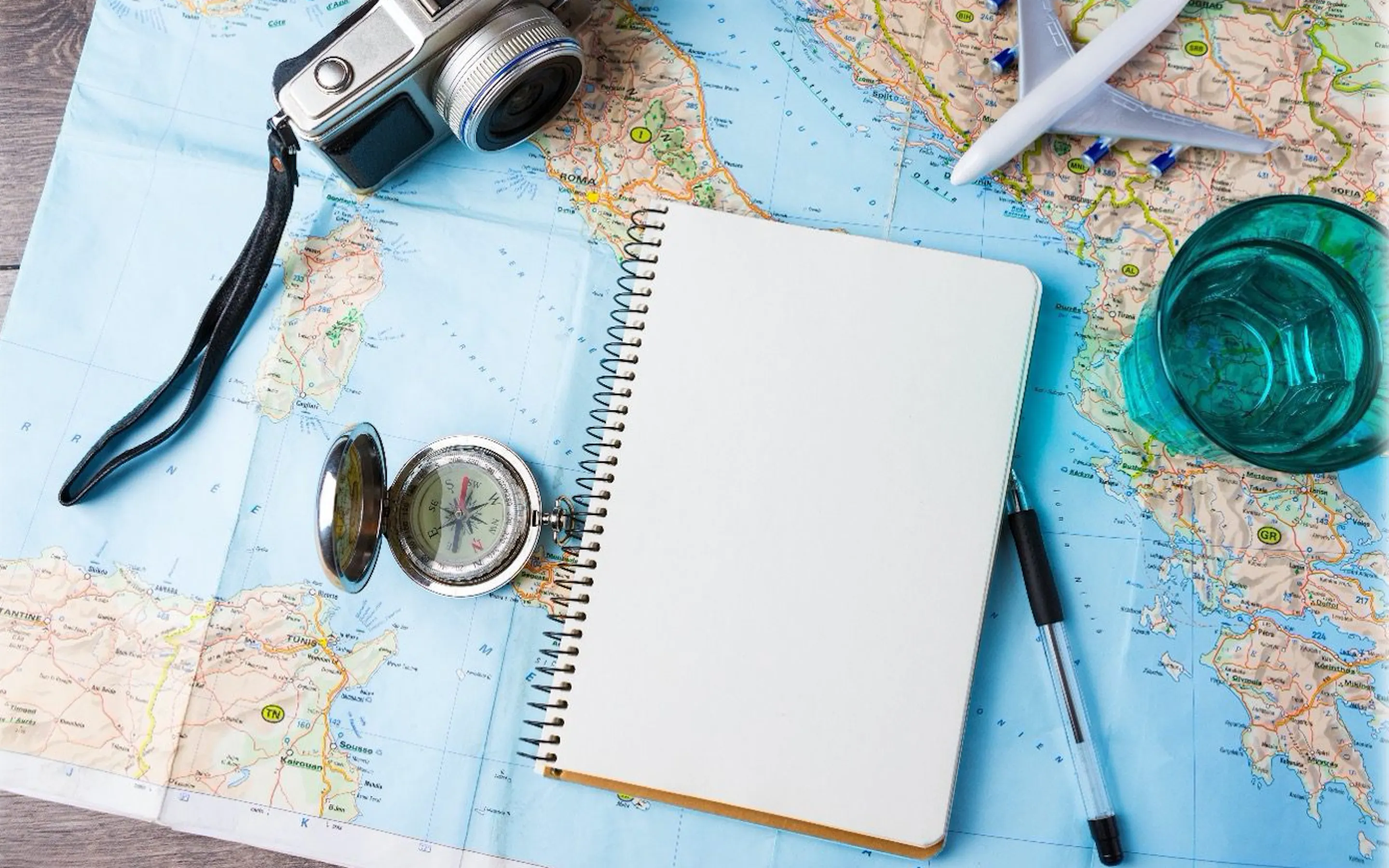 Travel flat lay with a map, notebook, passport and sunglasses, plus a camera, pen and drink on a white table.