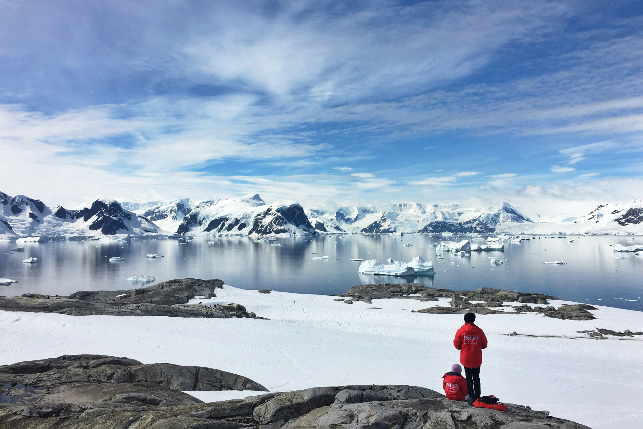 People in a red jackets stand and sit on a rocky Antarctic shore facing icebergs and snowy mountains across calm water.