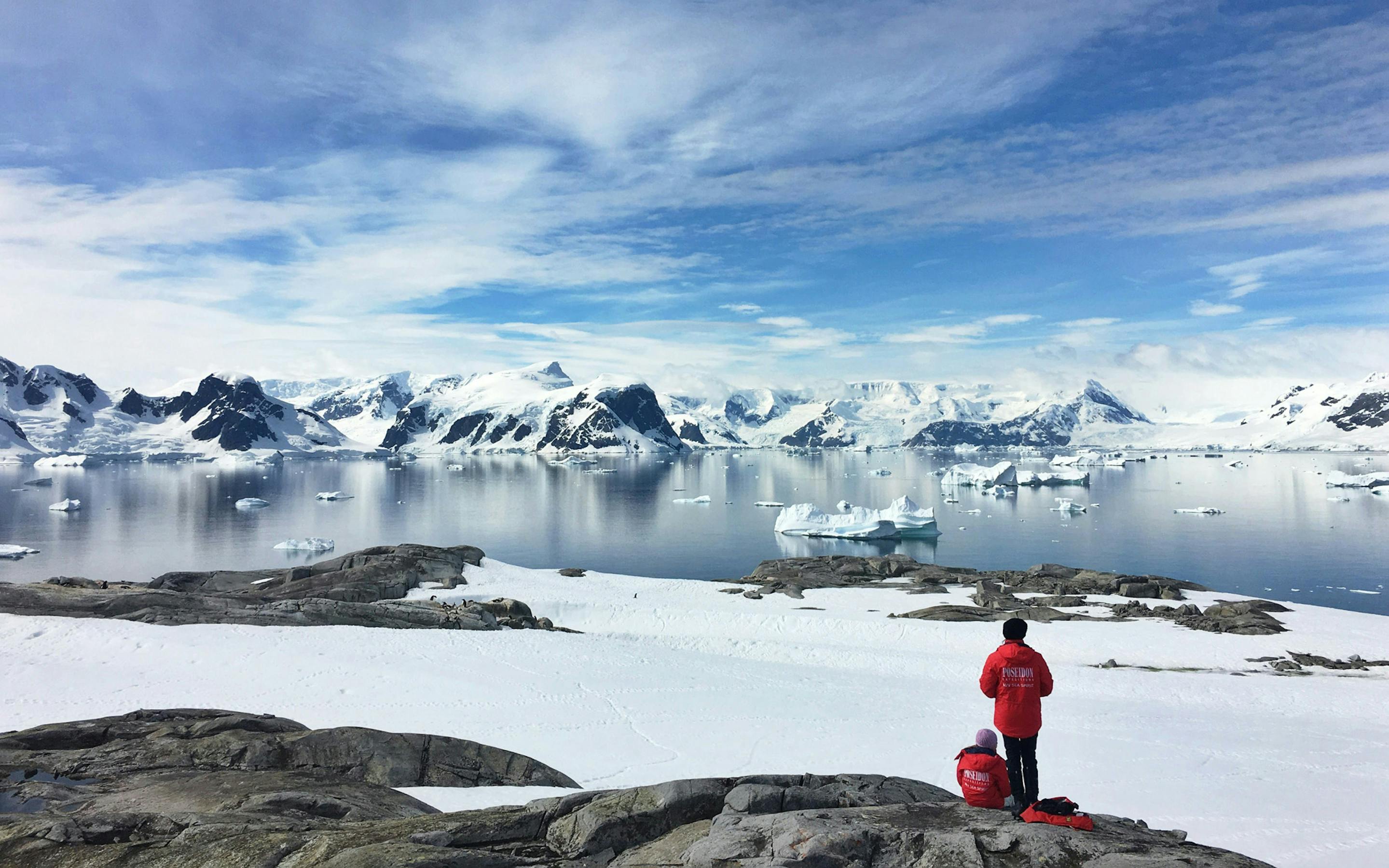 People in a red jackets stand and sit on a rocky Antarctic shore facing icebergs and snowy mountains across calm water.