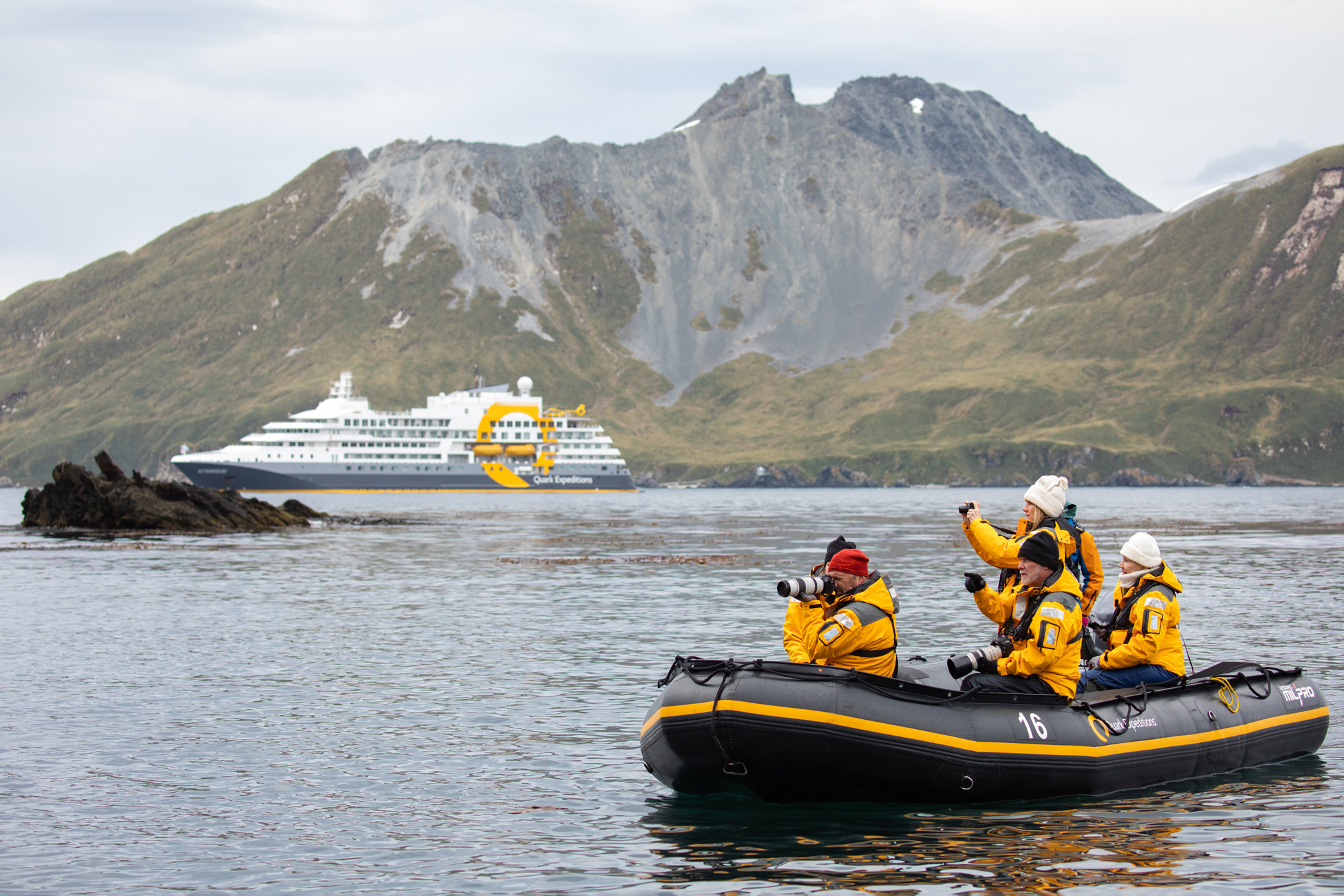 Passengers in a black and yellow zodiac boat approach the expedition ship Ultramarine near a rugged Antarctic coastline.