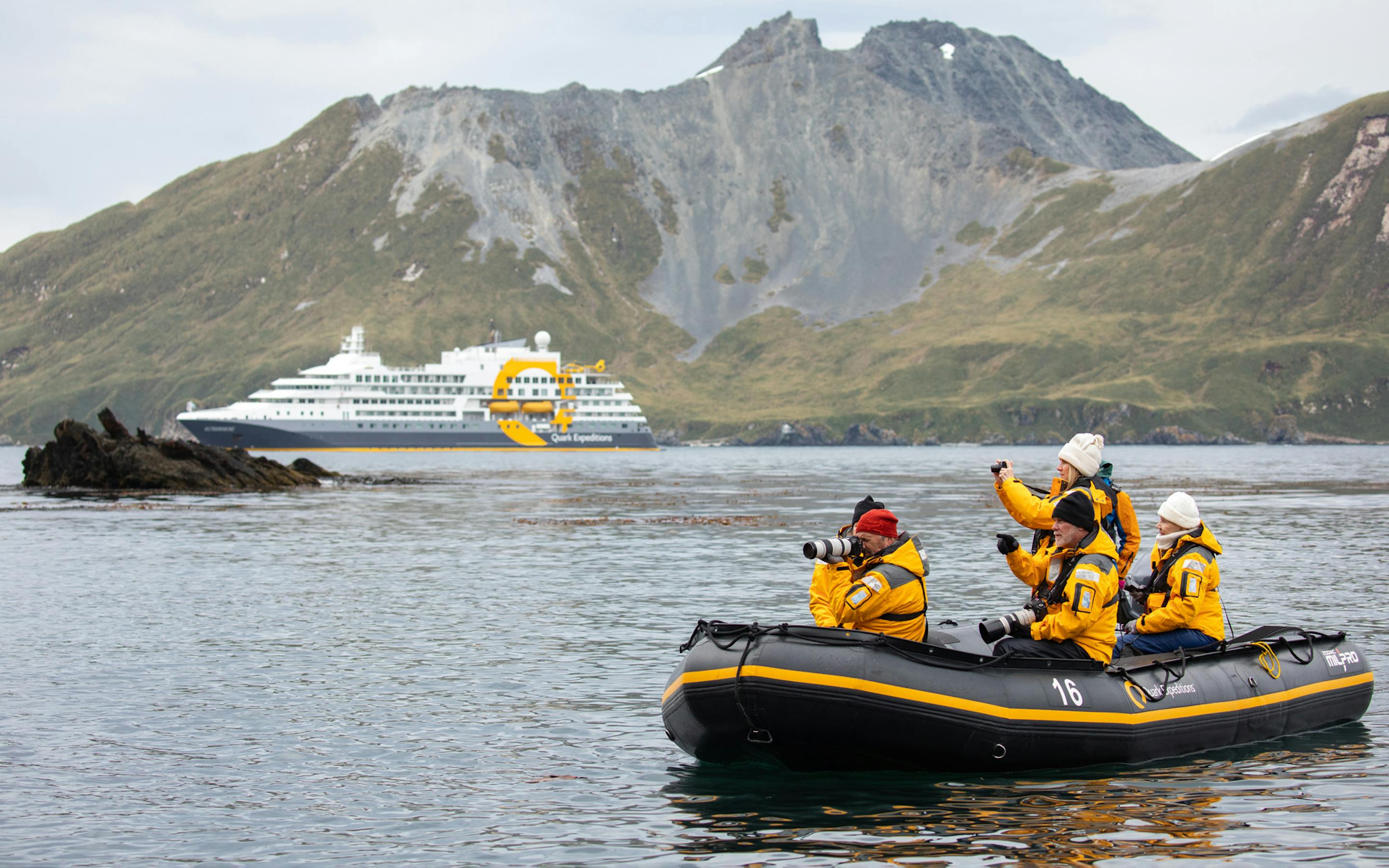 Passengers in a black and yellow zodiac boat approach the expedition ship Ultramarine near a rugged Antarctic coastline.
