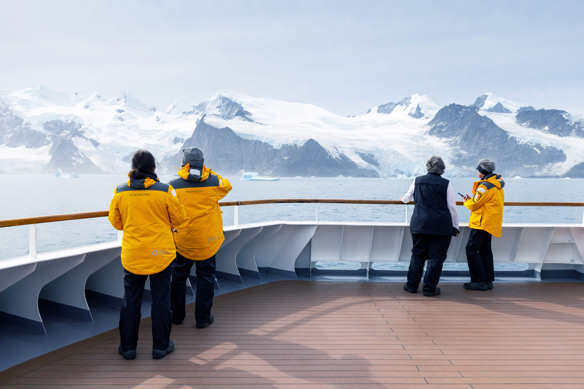 Passengers in yellow jackets watch Antarctic peaks from a ship’s deck rail, with icy water and clouds beyond.