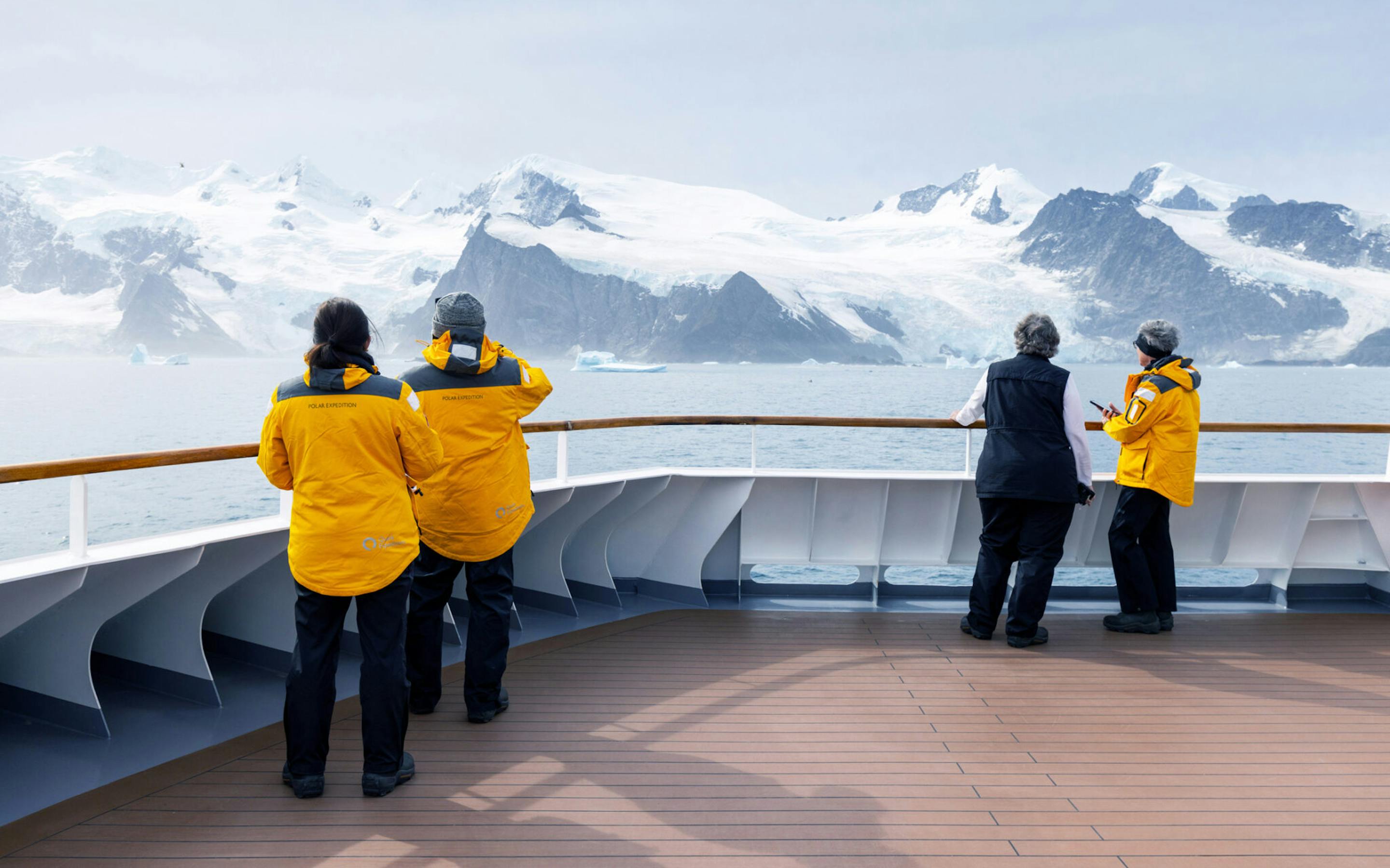 Passengers in yellow jackets watch Antarctic peaks from a ship’s deck rail, with icy water and clouds beyond.