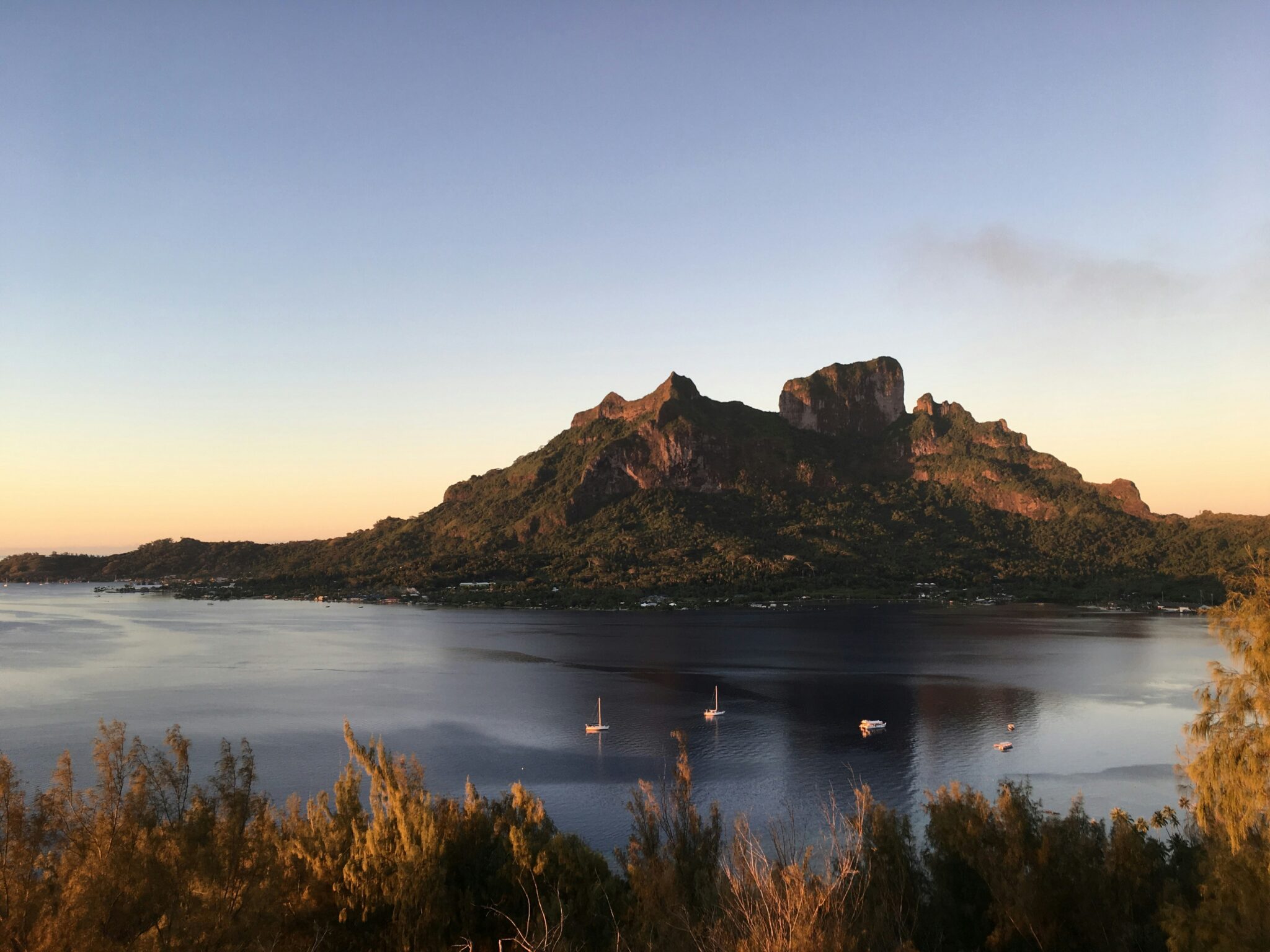 Mountain rising dramatically from a tranquil lagoon at sunset, with a few sailboats anchored in calm water and warm golden light illuminating the landscape.
