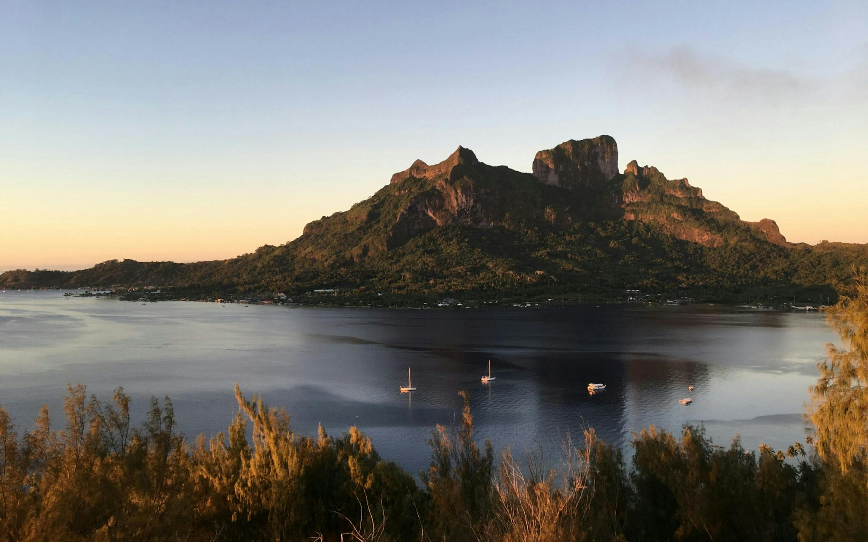 Mountain rising dramatically from a tranquil lagoon at sunset, with a few sailboats anchored in calm water and warm golden light illuminating the landscape.