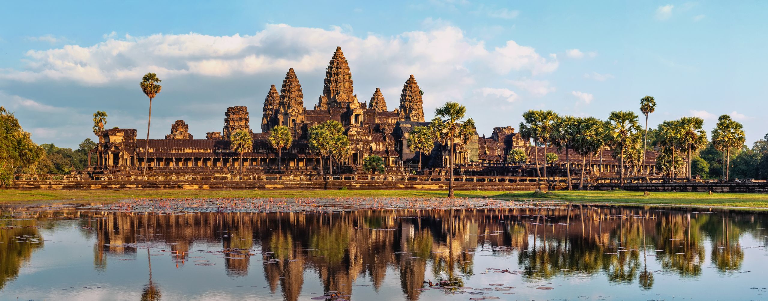 Angkor Wat temple complex reflected in a calm pond at sunrise, with palm trees lining the ancient stone structures under a soft blue sky.