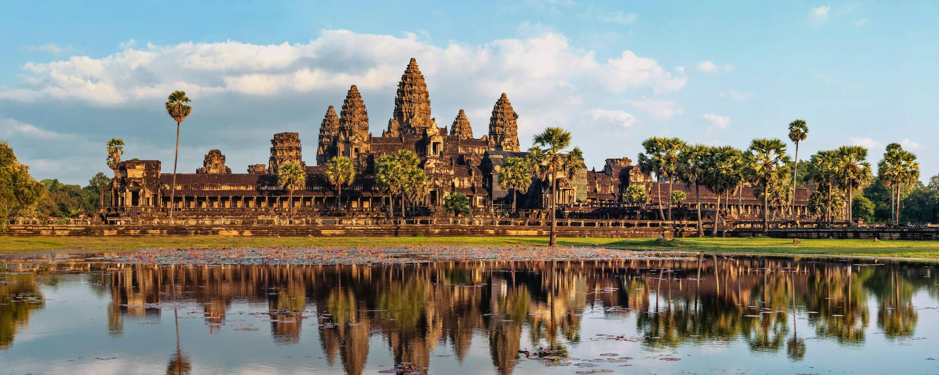 Angkor Wat temple complex reflected in a calm pond at sunrise, with palm trees lining the ancient stone structures under a soft blue sky.
