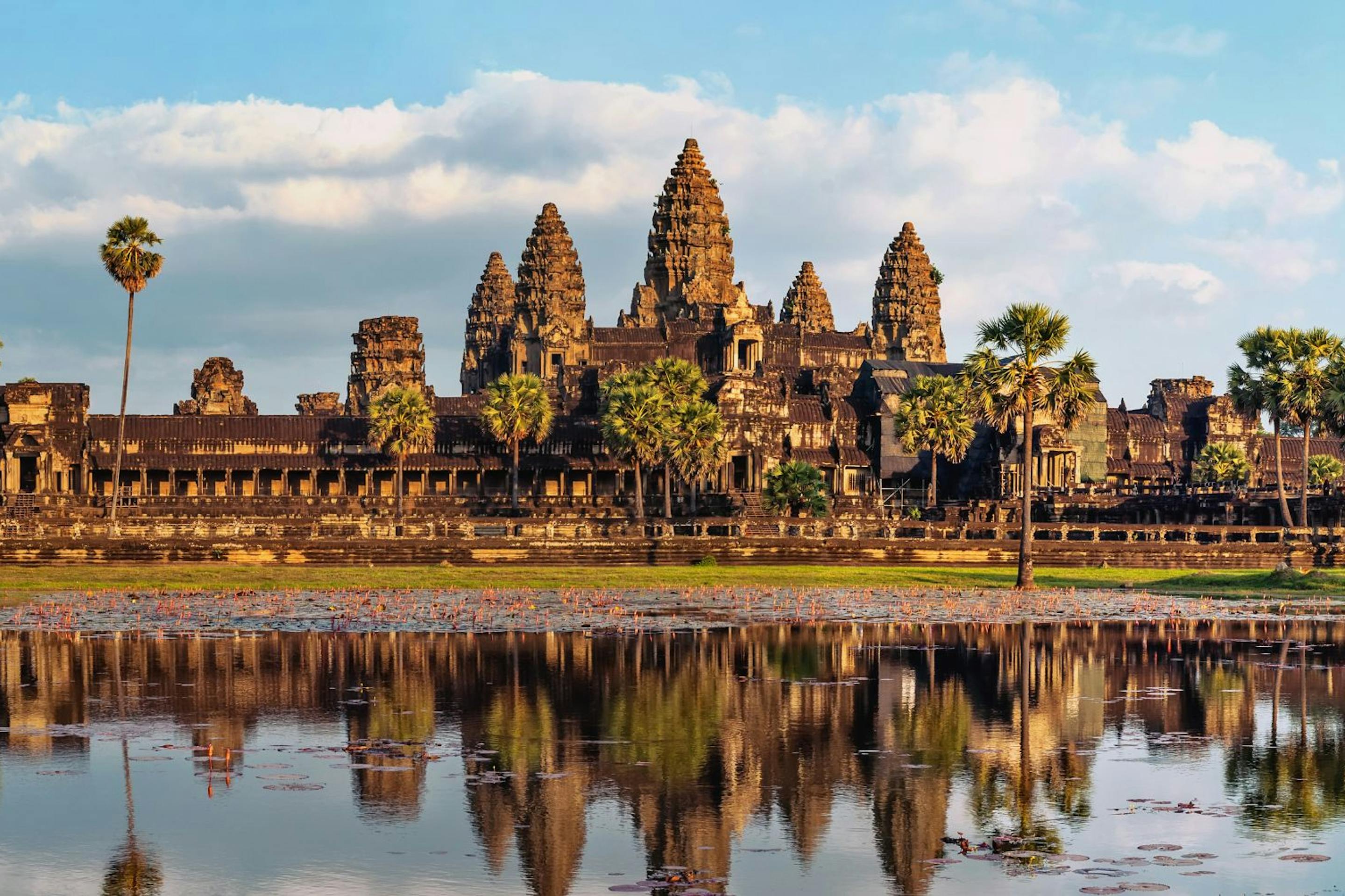 Angkor Wat temple complex reflected in a calm pond at sunrise, with palm trees lining the ancient stone structures under a soft blue sky.