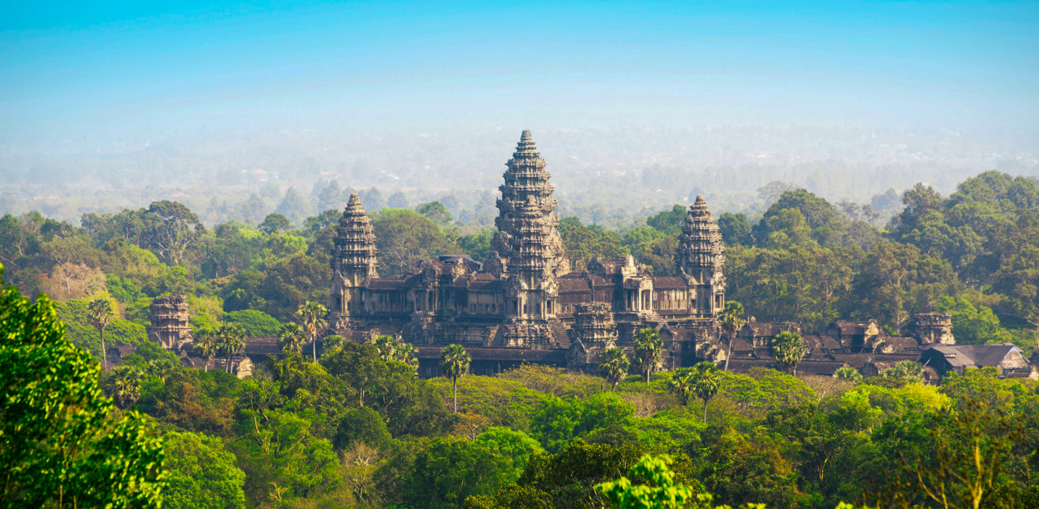 Angkor Wat rising above lush green forest canopy, its iconic central towers visible against a hazy blue sky.