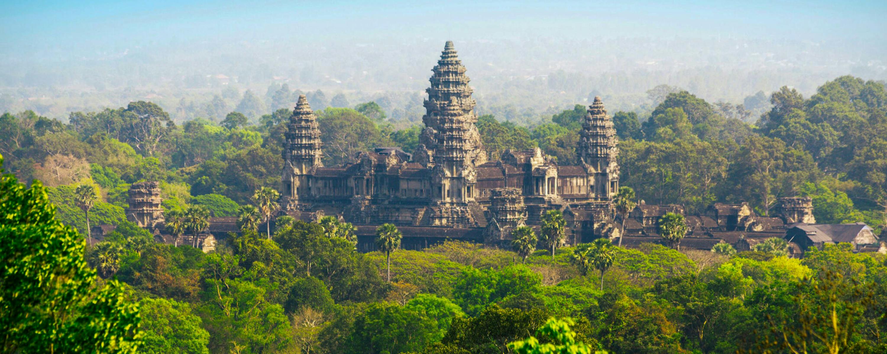Angkor Wat rising above lush green forest canopy, its iconic central towers visible against a hazy blue sky.
