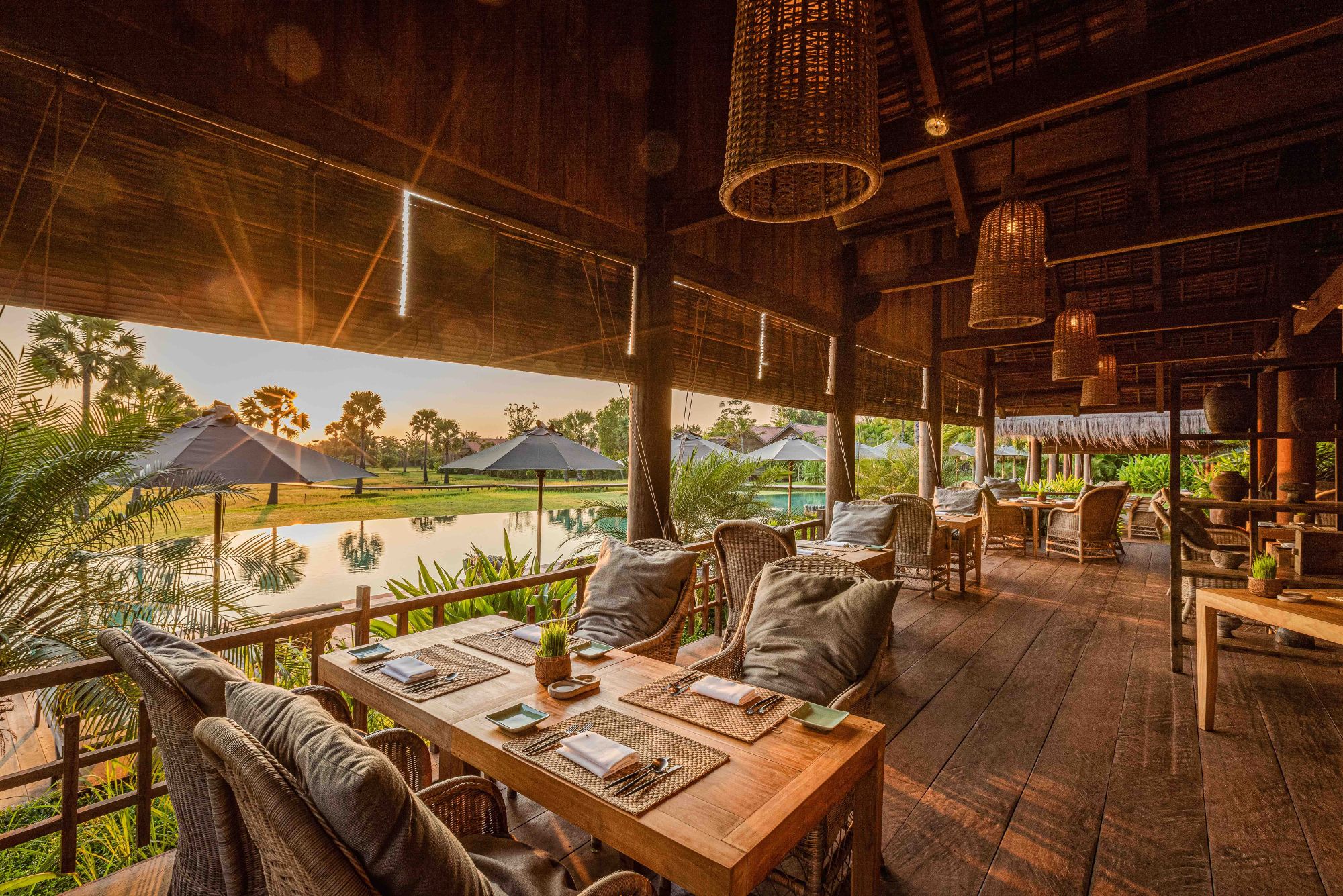 Open-air wooden terrace restaurant overlooking a tranquil pool and gardens at sunset, with woven lanterns and neatly set dining tables.