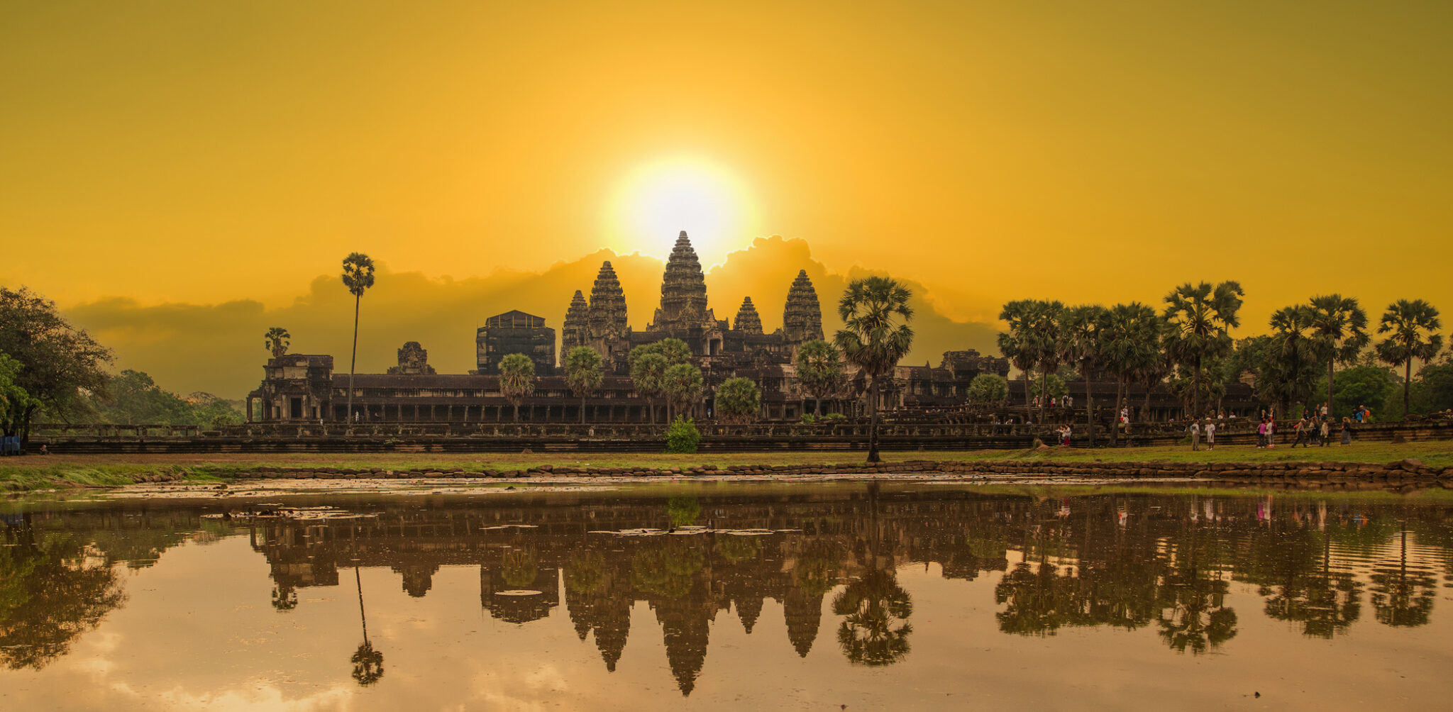 Angkor Wat silhouetted against a vibrant golden sunrise, reflected in a still pond with palm trees framing the ancient temple.