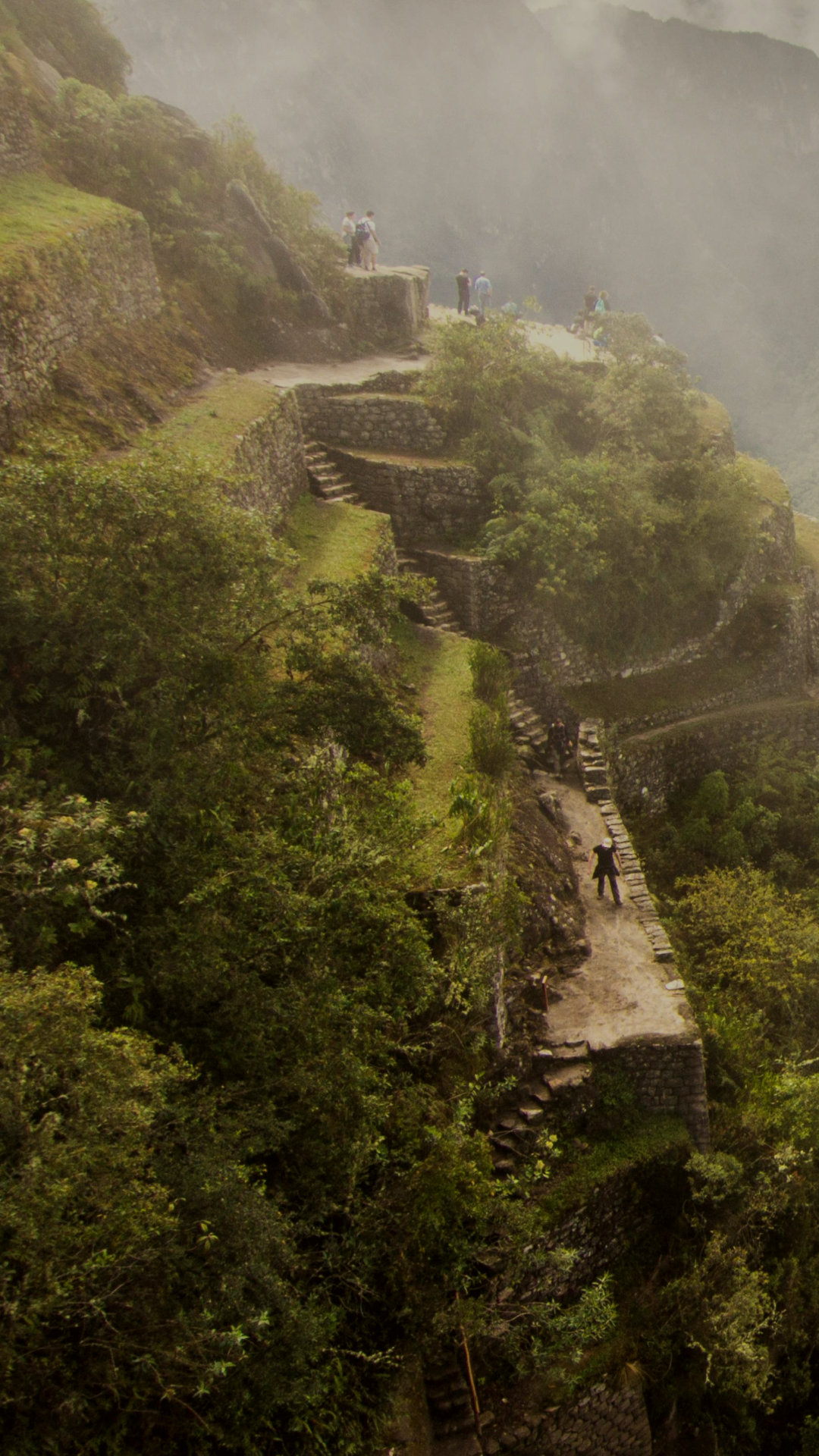 Stone terraces and narrow pathways winding along a steep, misty mountainside, with visitors exploring the ancient ruins surrounded by lush green vegetation.