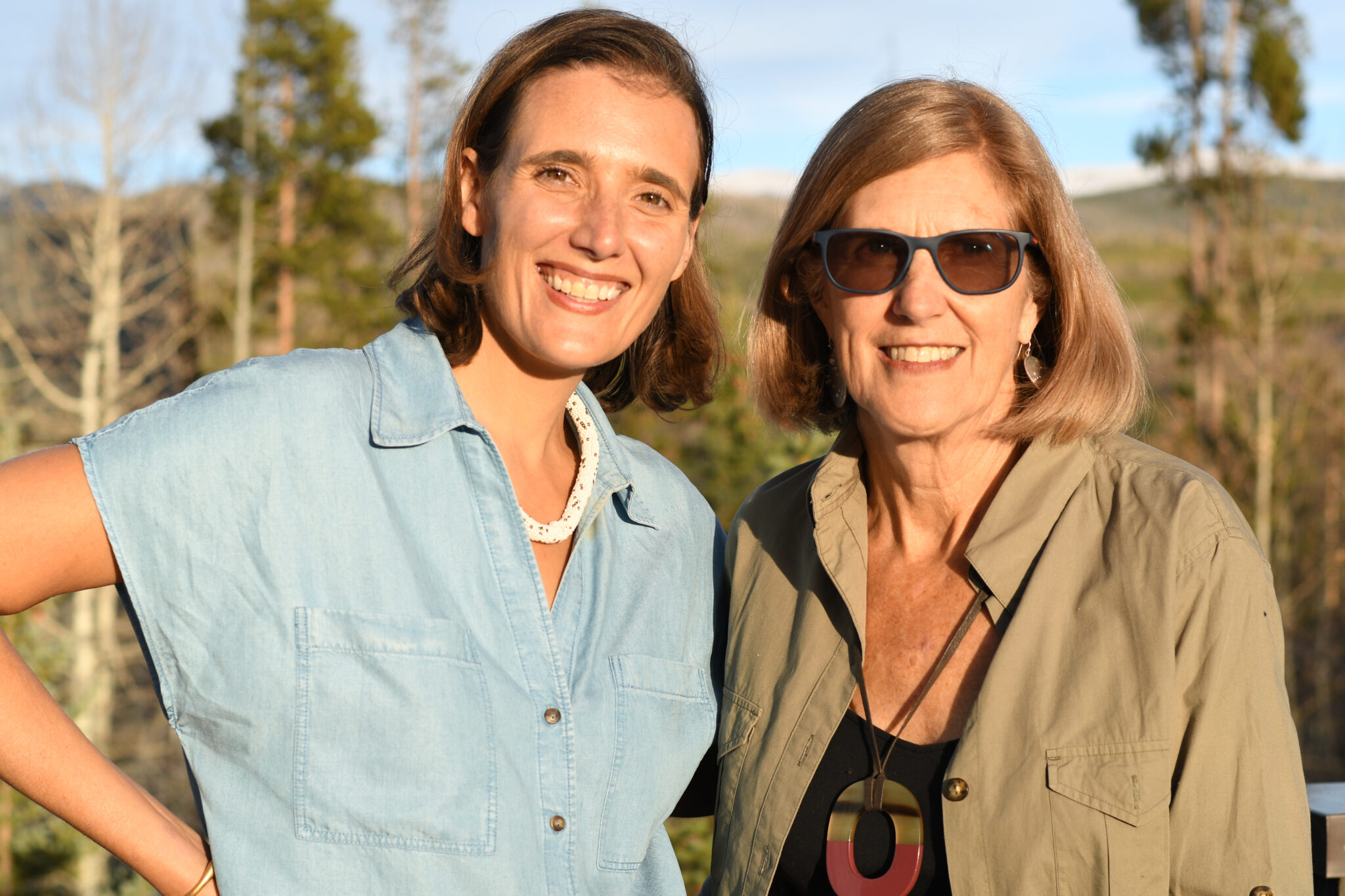 Two women smiling outdoors in warm sunlight, standing close together with trees and hills in the background.