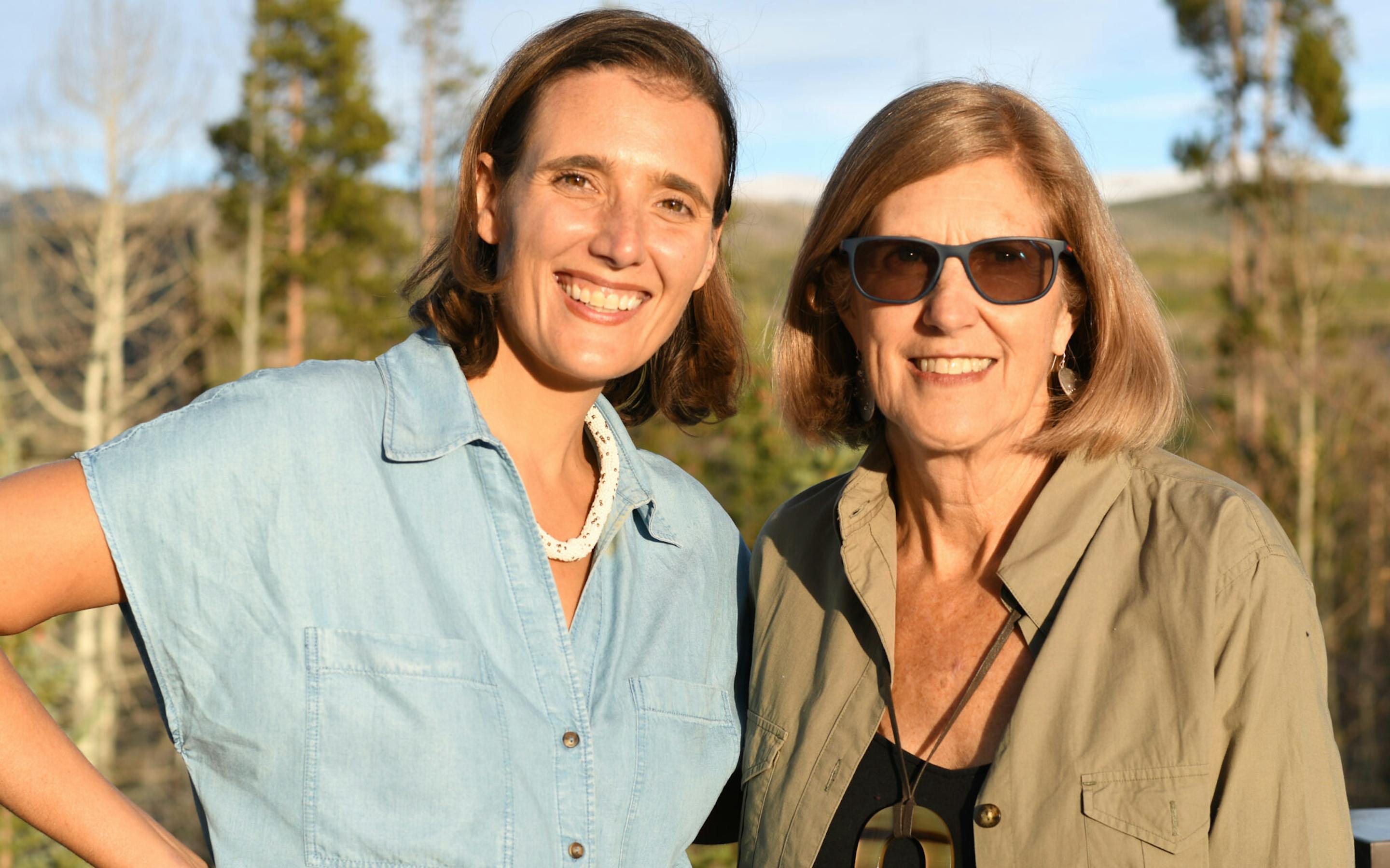 Two women smiling outdoors in warm sunlight, standing close together with trees and hills in the background.