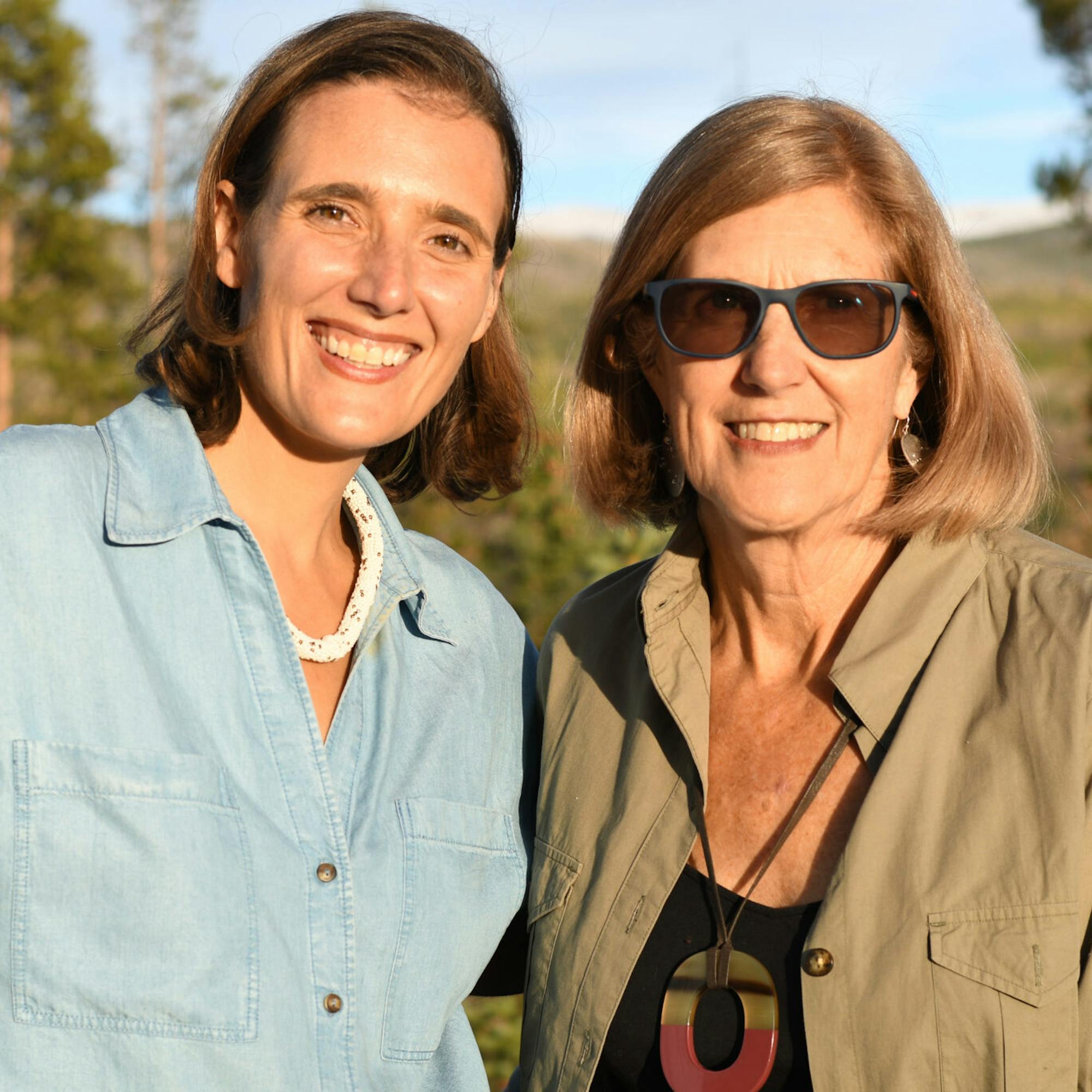 Two women smiling outdoors in warm sunlight, standing close together with trees and hills in the background.