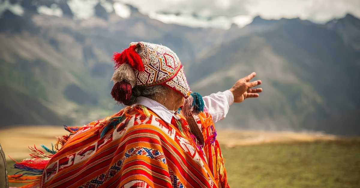 Person in traditional Andean clothing with a colorful woven poncho and hat, gesturing toward dramatic mountain peaks in the background.