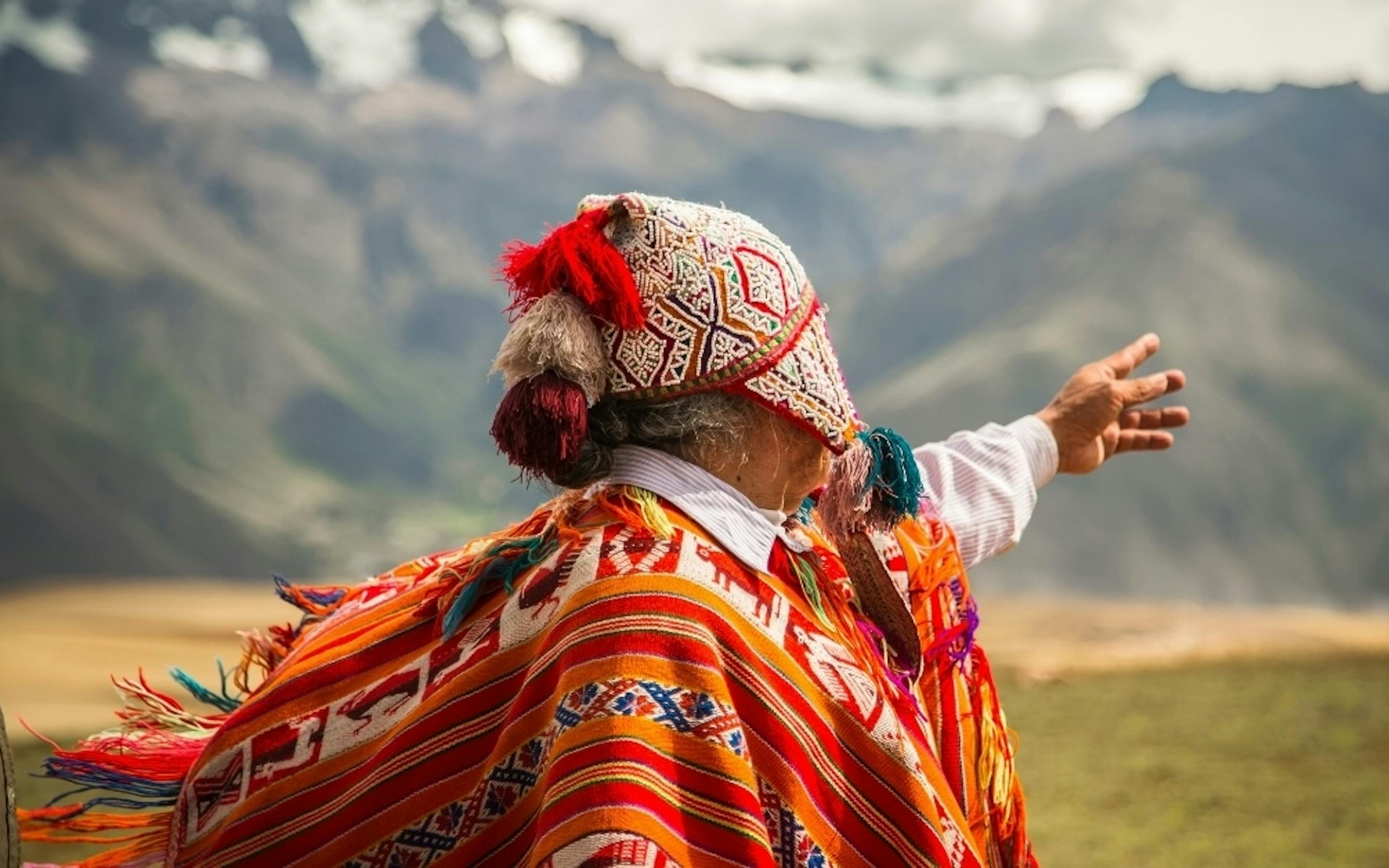 Person in traditional Andean clothing with a colorful woven poncho and hat, gesturing toward dramatic mountain peaks in the background.
