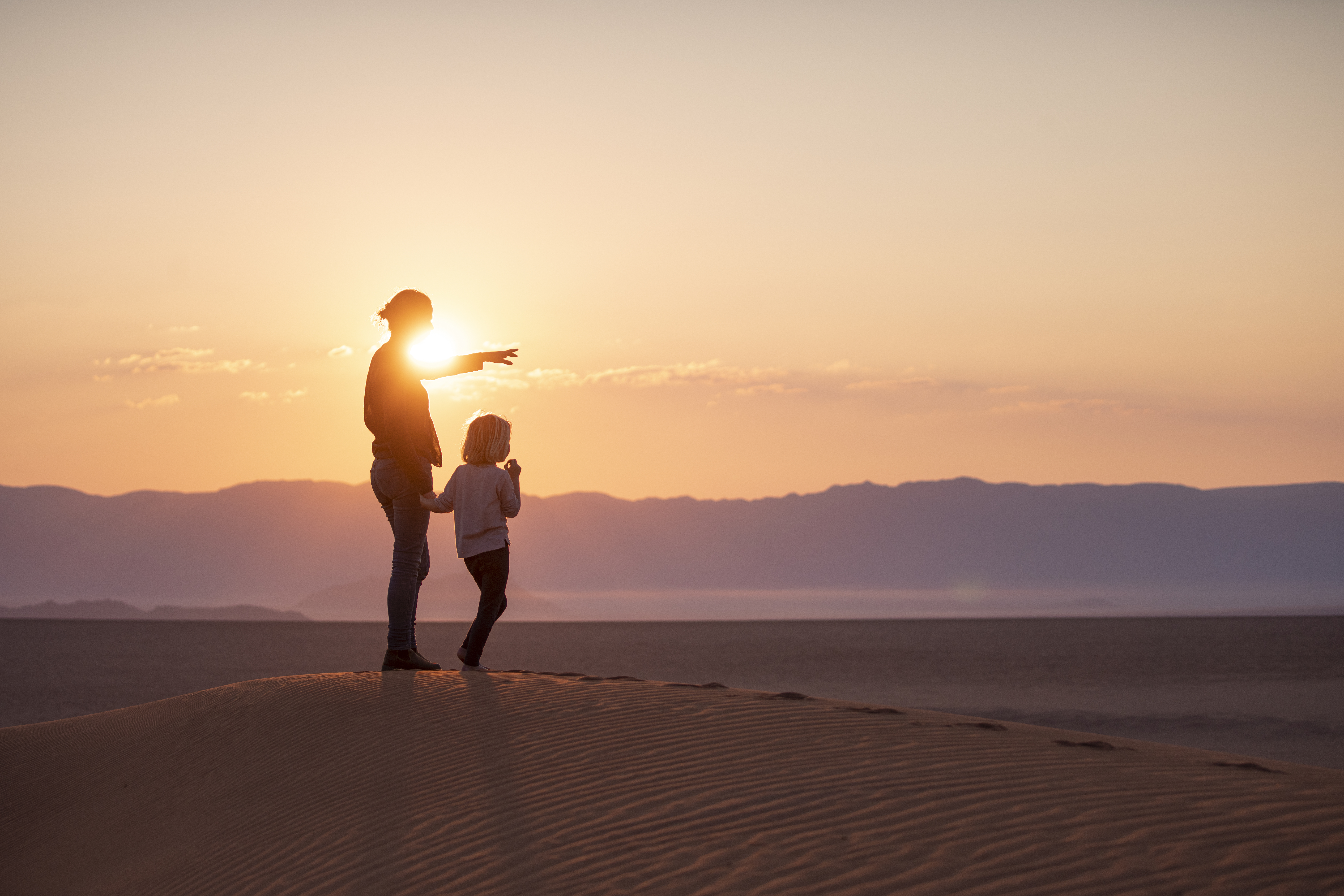 Silhouette of an adult and child standing on a sand dune at sunset, pointing toward distant mountains beneath a glowing orange sky.