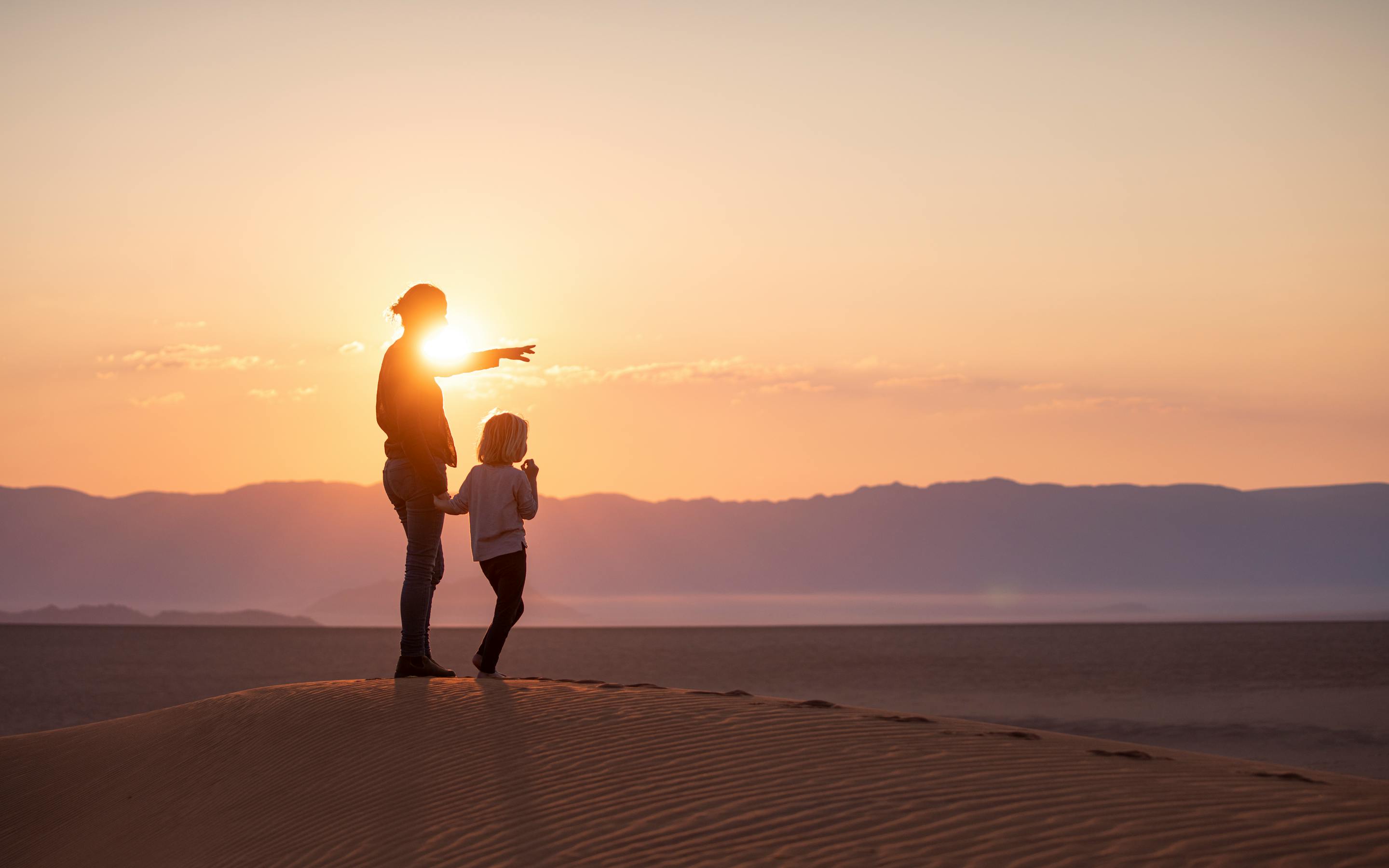 Silhouette of an adult and child standing on a sand dune at sunset, pointing toward distant mountains beneath a glowing orange sky.
