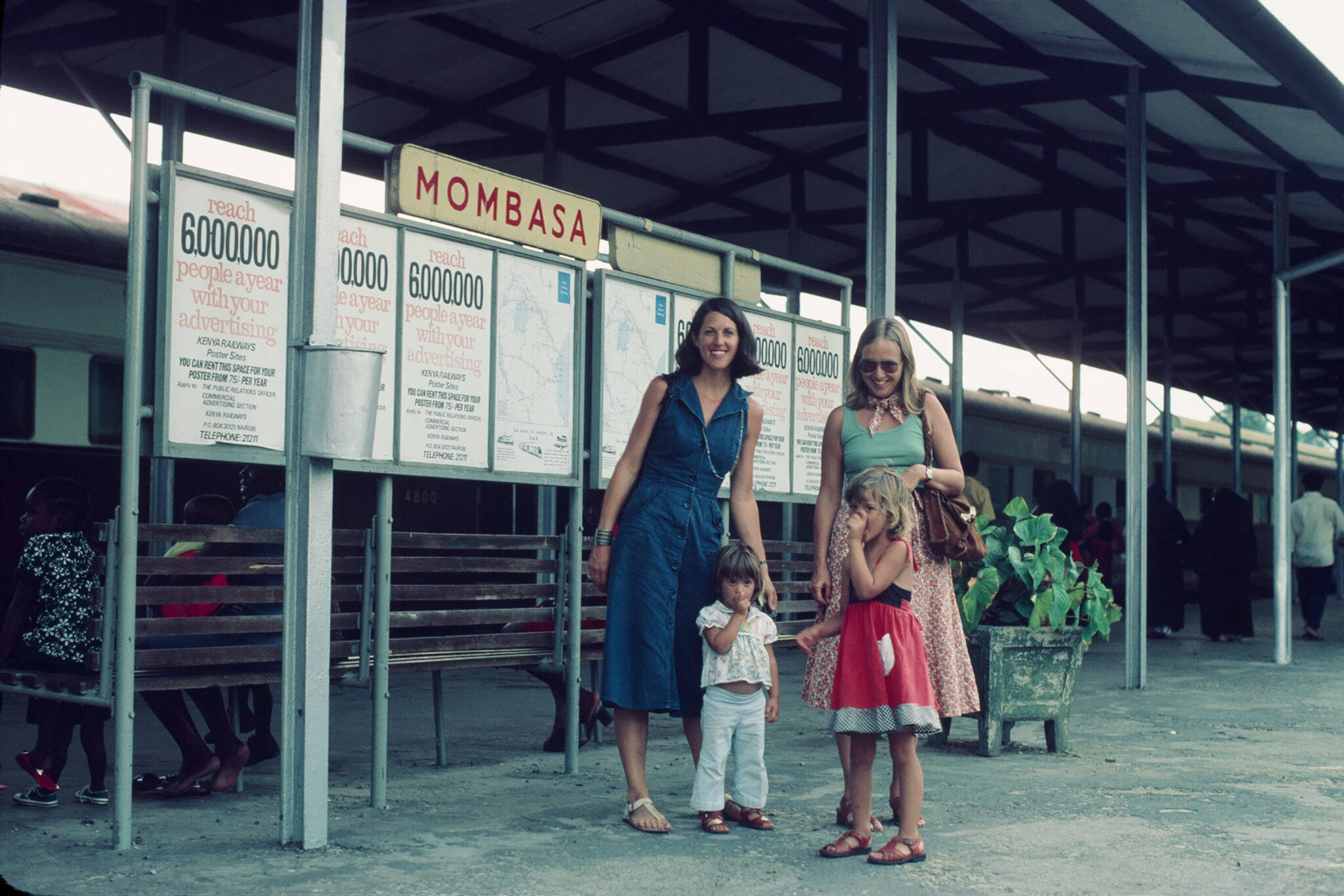 Two women and two young children standing on a train platform beneath a “Mombasa” sign, with a passenger train and posted advertisements in the background.