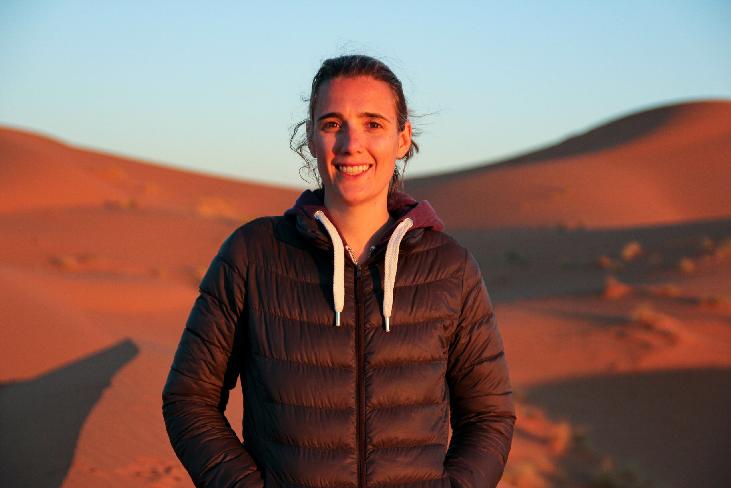 Woman in a dark jacket standing on red sand dunes at sunset, illuminated by warm golden light.