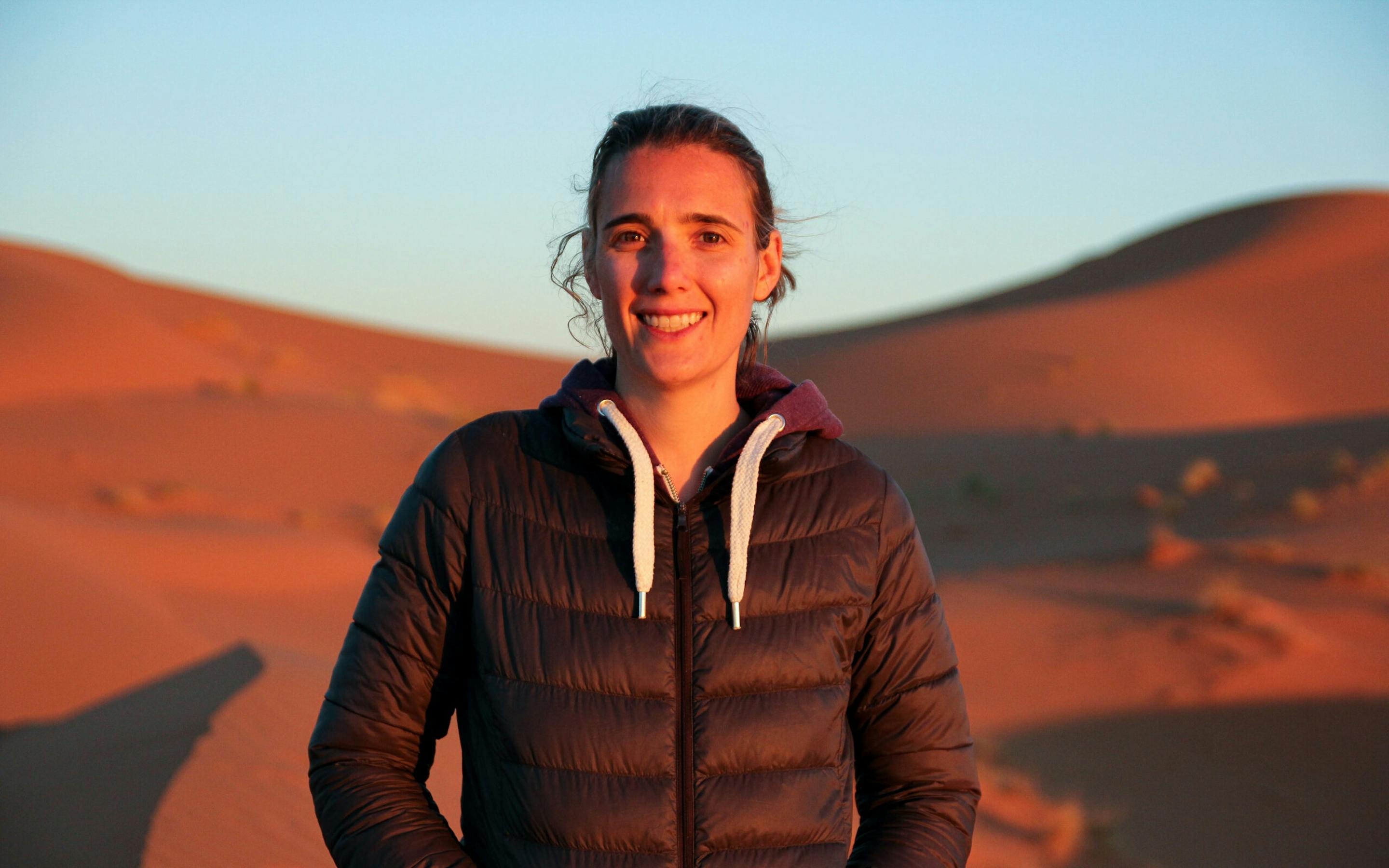 Woman in a dark jacket standing on red sand dunes at sunset, illuminated by warm golden light.