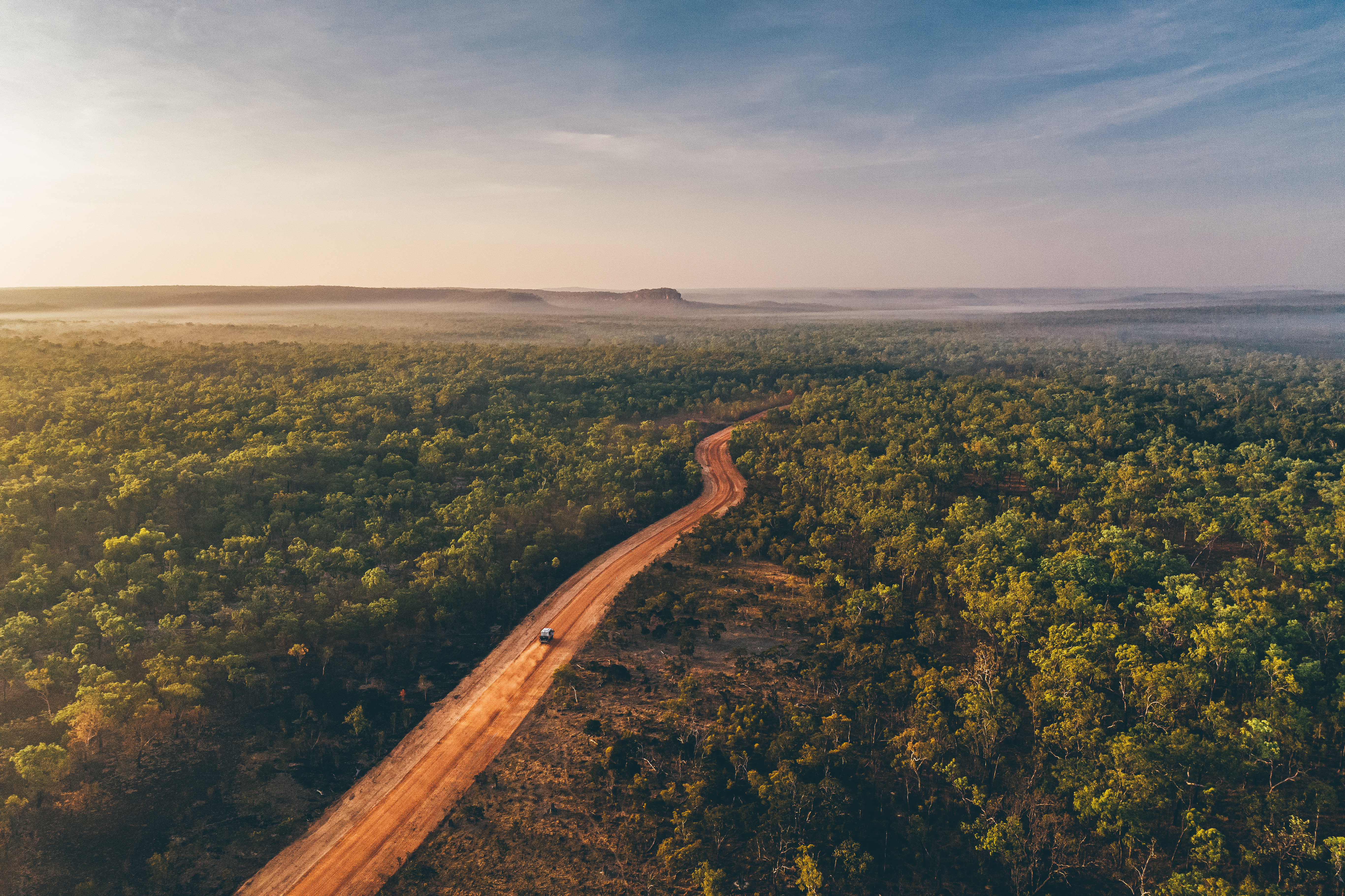 An aerial view shows a red dirt road cutting through dense green forest, fading into haze at the horizon.