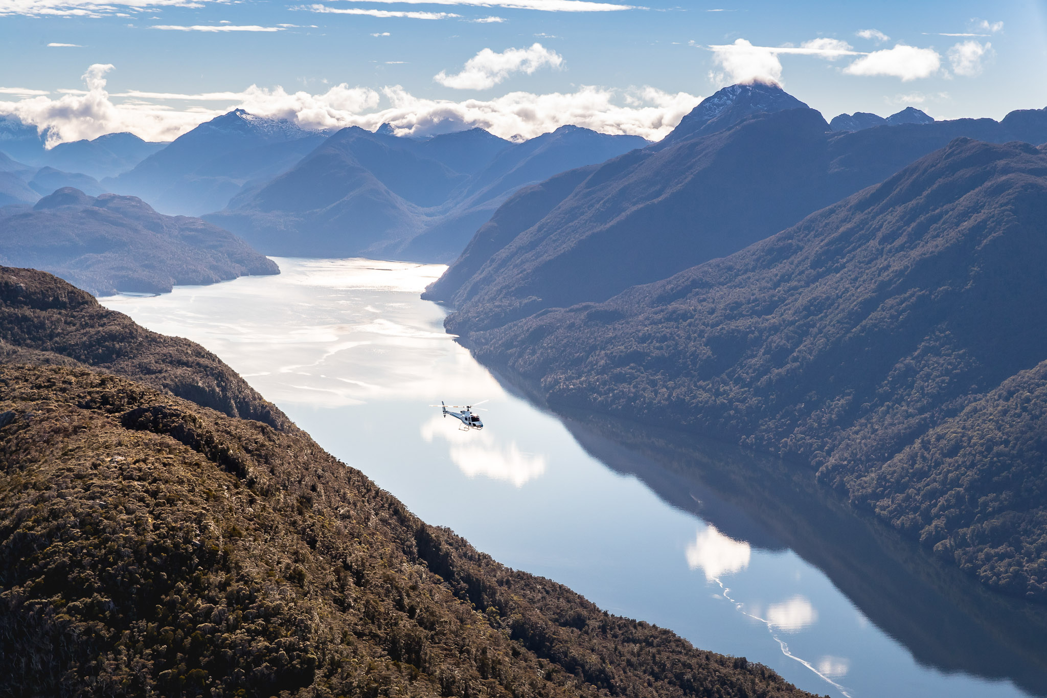 A winding alpine lake threads through steep ridgelines, its pale surface catching sun above shadowed valleys. A helicopter flies along the lake.