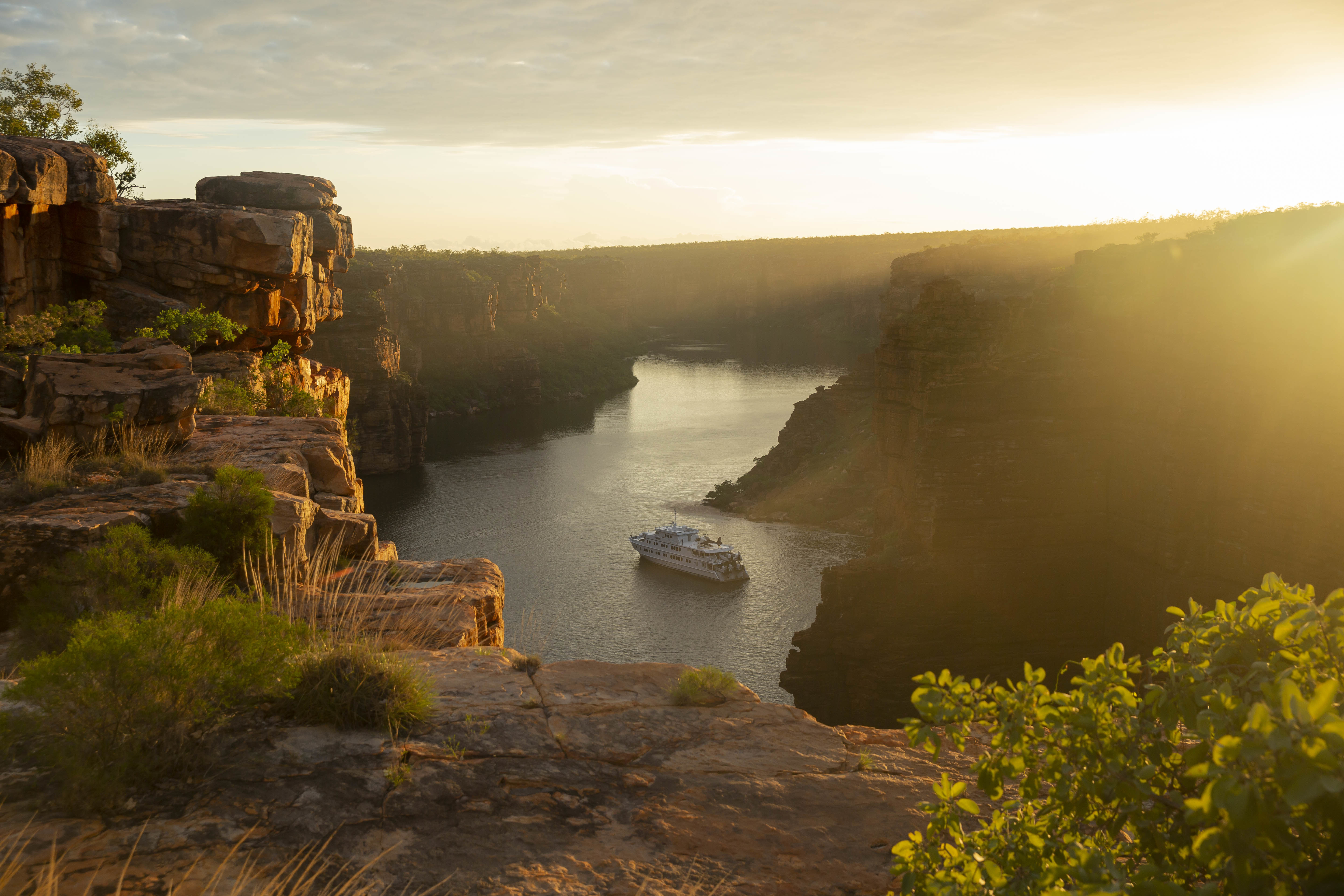 Golden sunset lights a winding river gorge, with rugged cliffs and a small boat traveling through the calm channel.
