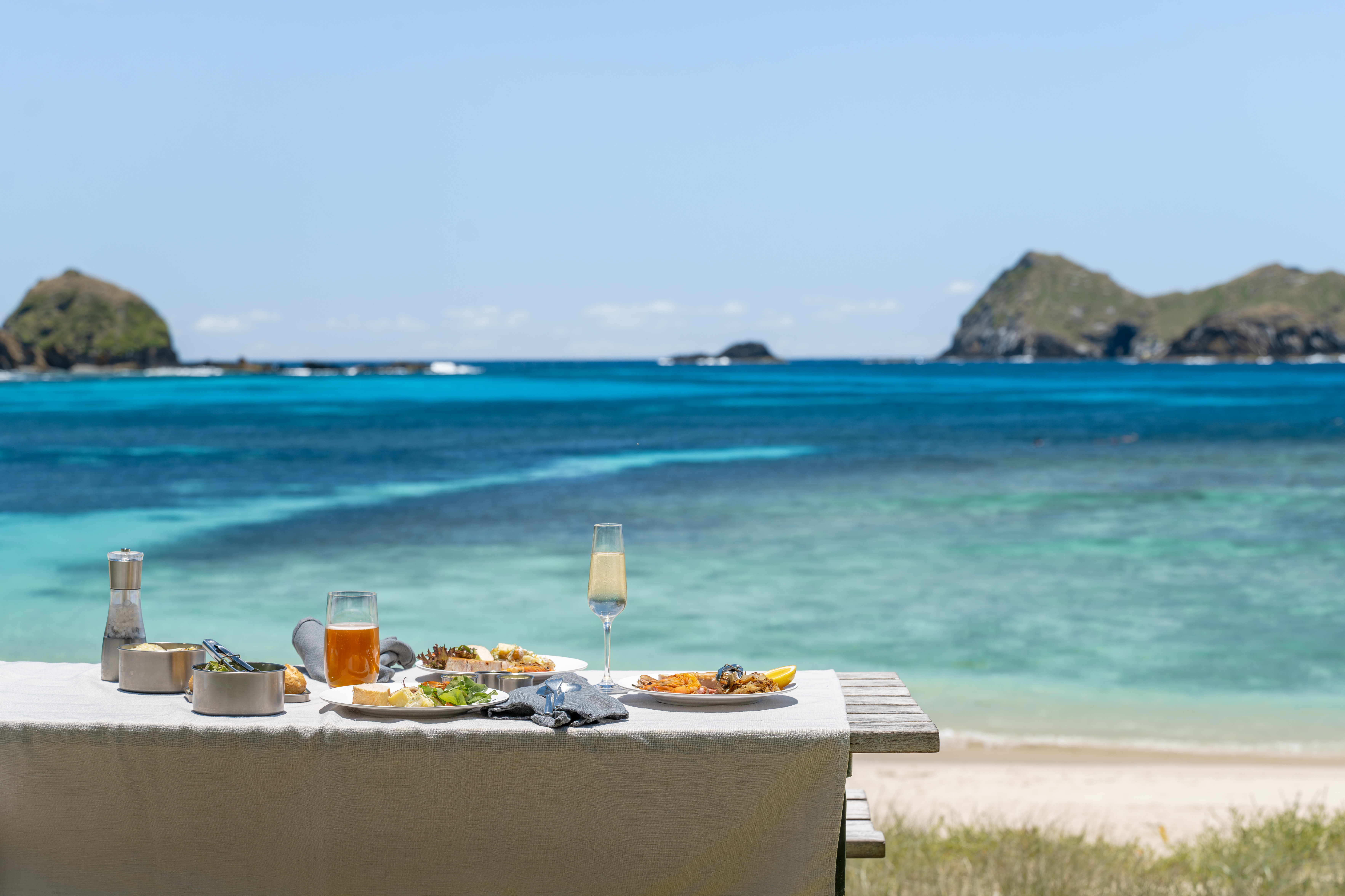 A picnic table sits above a turquoise lagoon, with two rugged islets offshore and gentle waves on the sand.