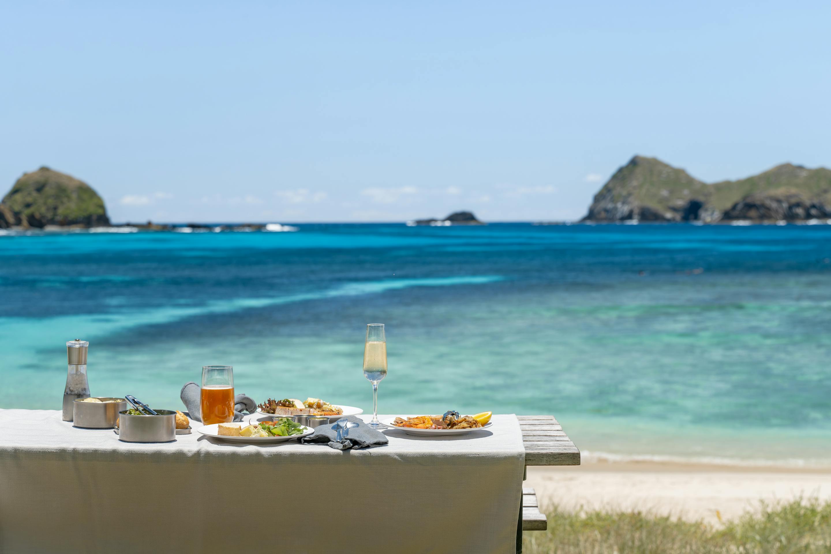A picnic table sits above a turquoise lagoon, with two rugged islets offshore and gentle waves on the sand.