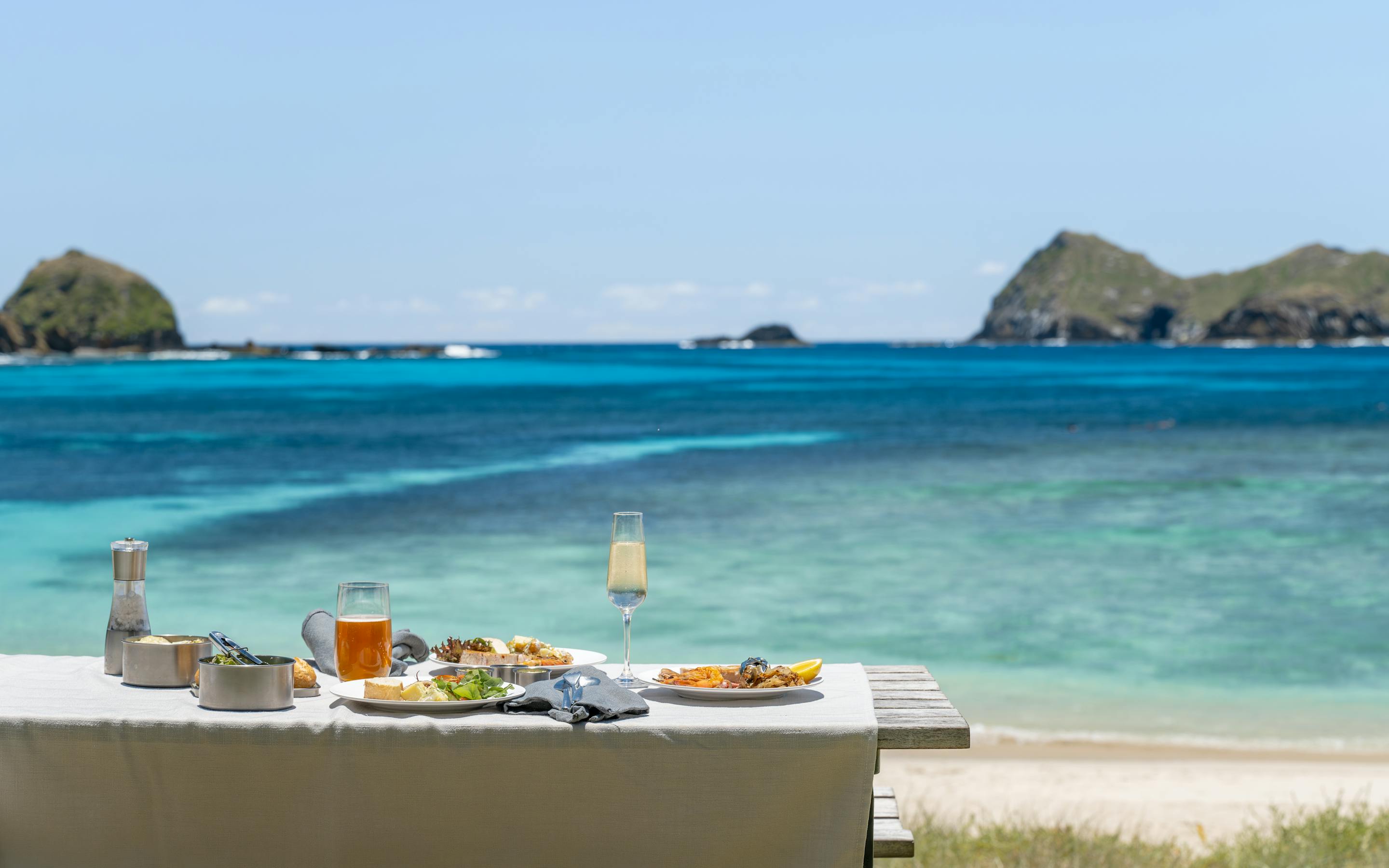 A picnic table sits above a turquoise lagoon, with two rugged islets offshore and gentle waves on the sand.