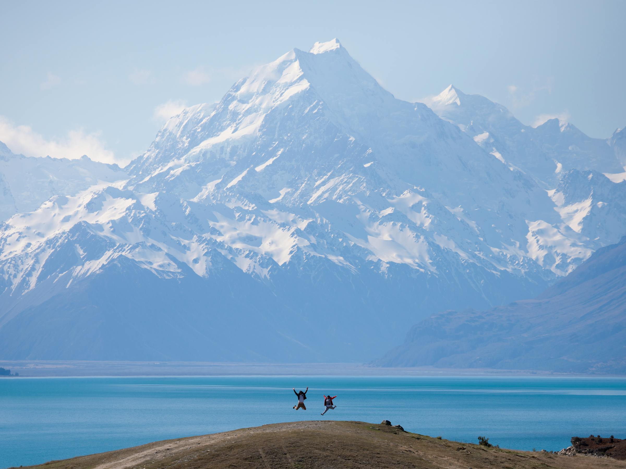 Two figures stand on a lakeshore lookout above blue Lake Pukaki, with snowcapped Aoraki rising beyond the water.