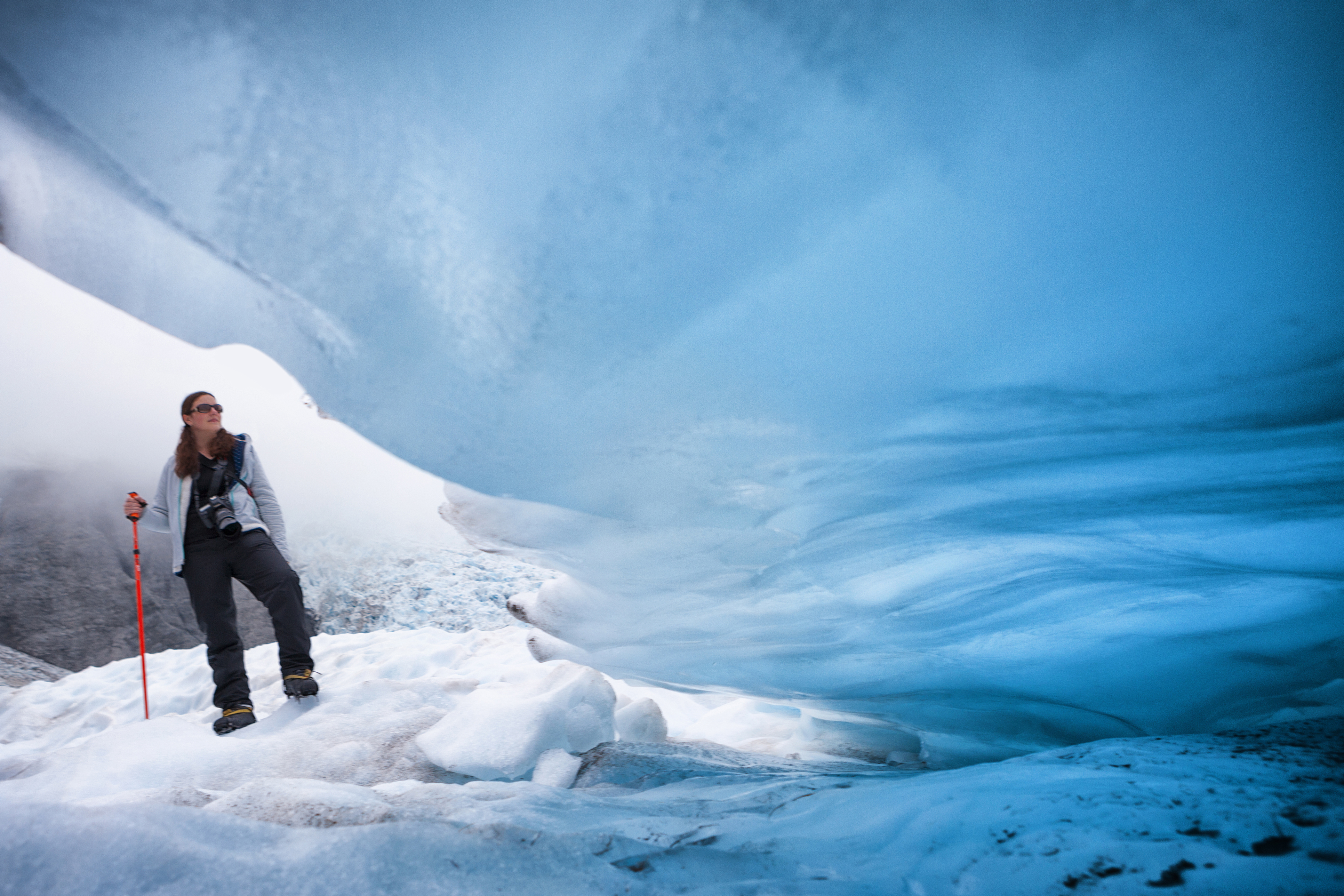 A hiker walks across blue glacial ice beside a cave opening, jagged frozen walls glowing in soft pale light.