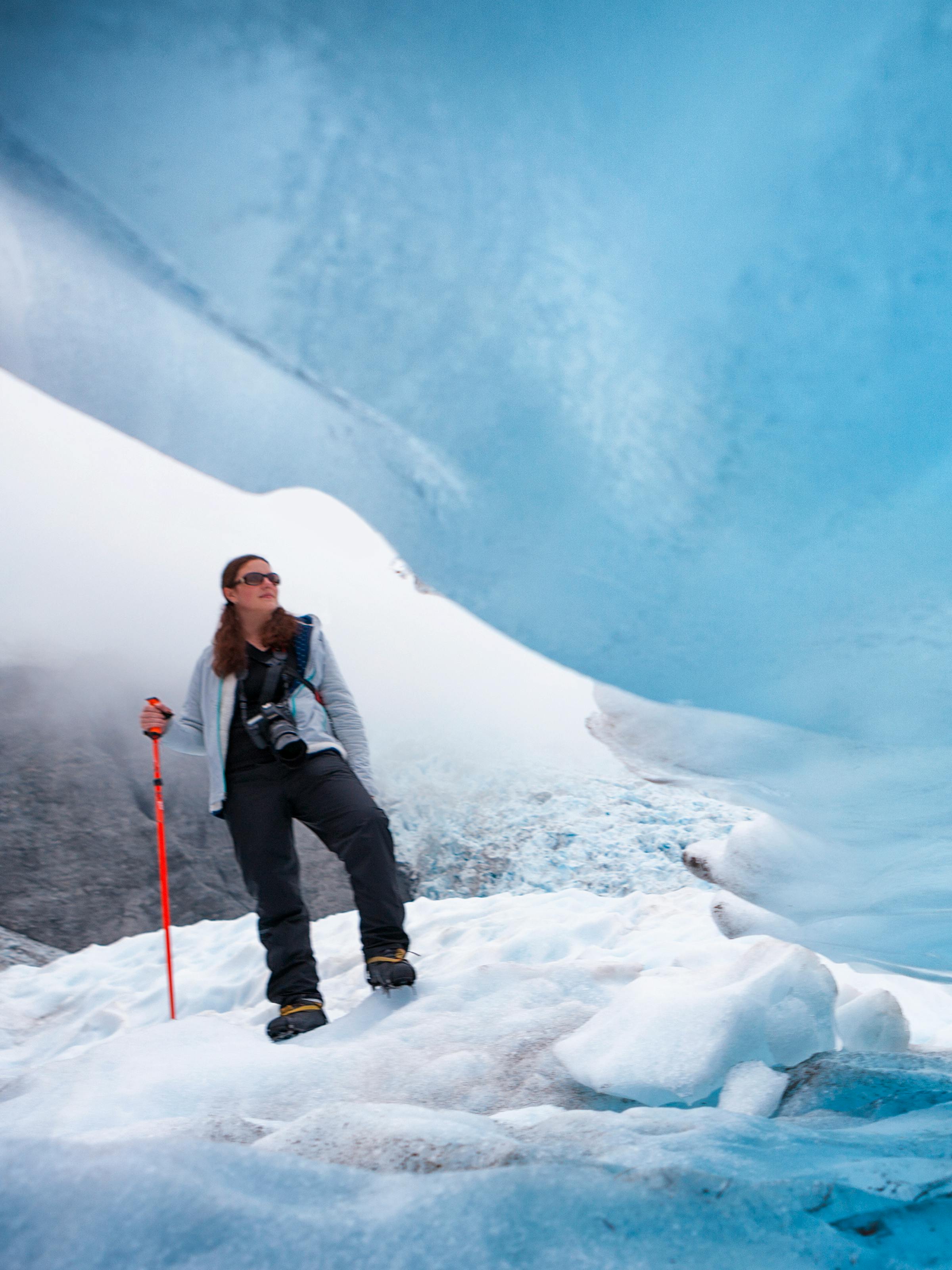 A hiker walks across blue glacial ice beside a cave opening, jagged frozen walls glowing in soft pale light.