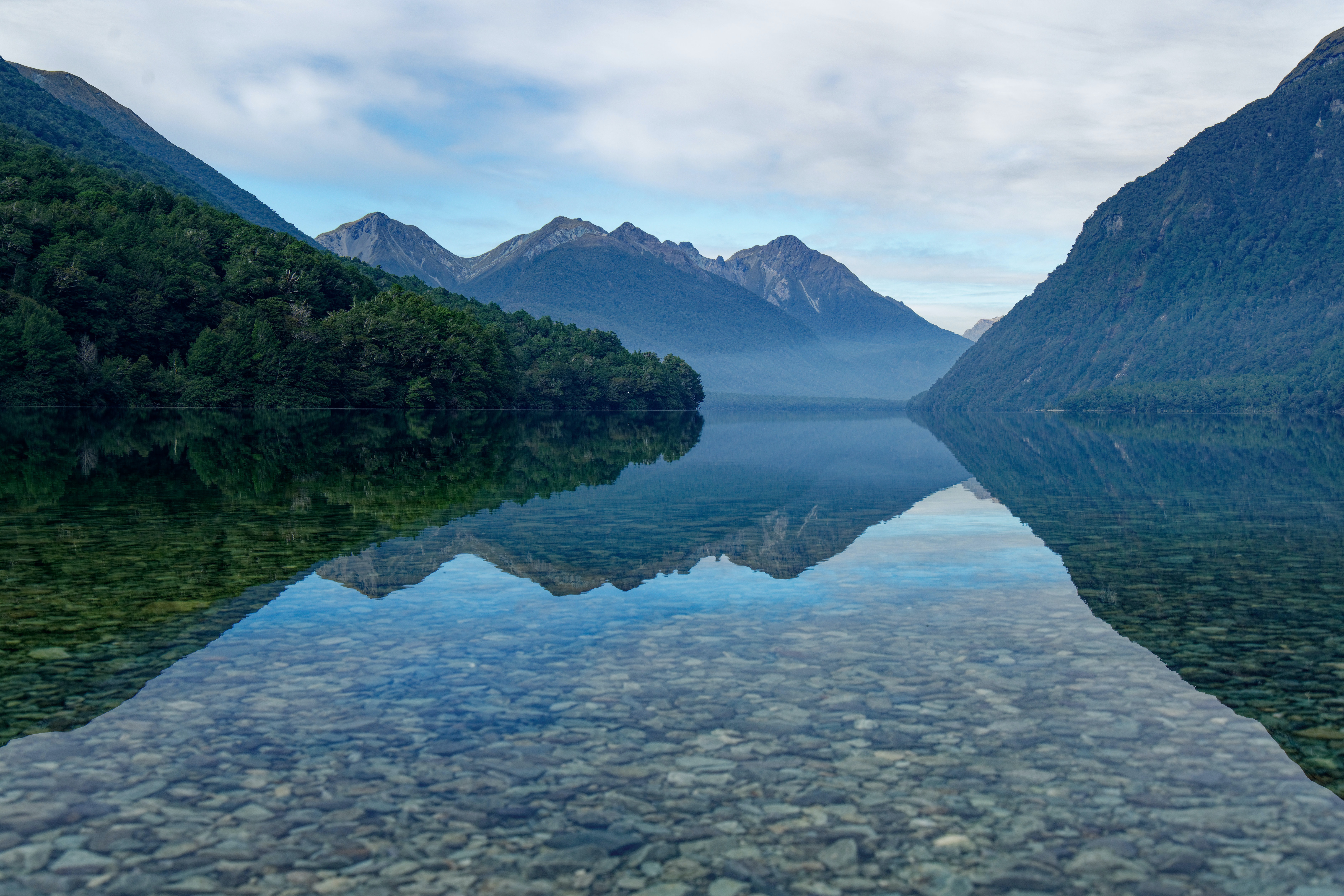 Still water at Gun Lake reflects forested slopes and misty peaks, the rocky shore visible through clear shallows.