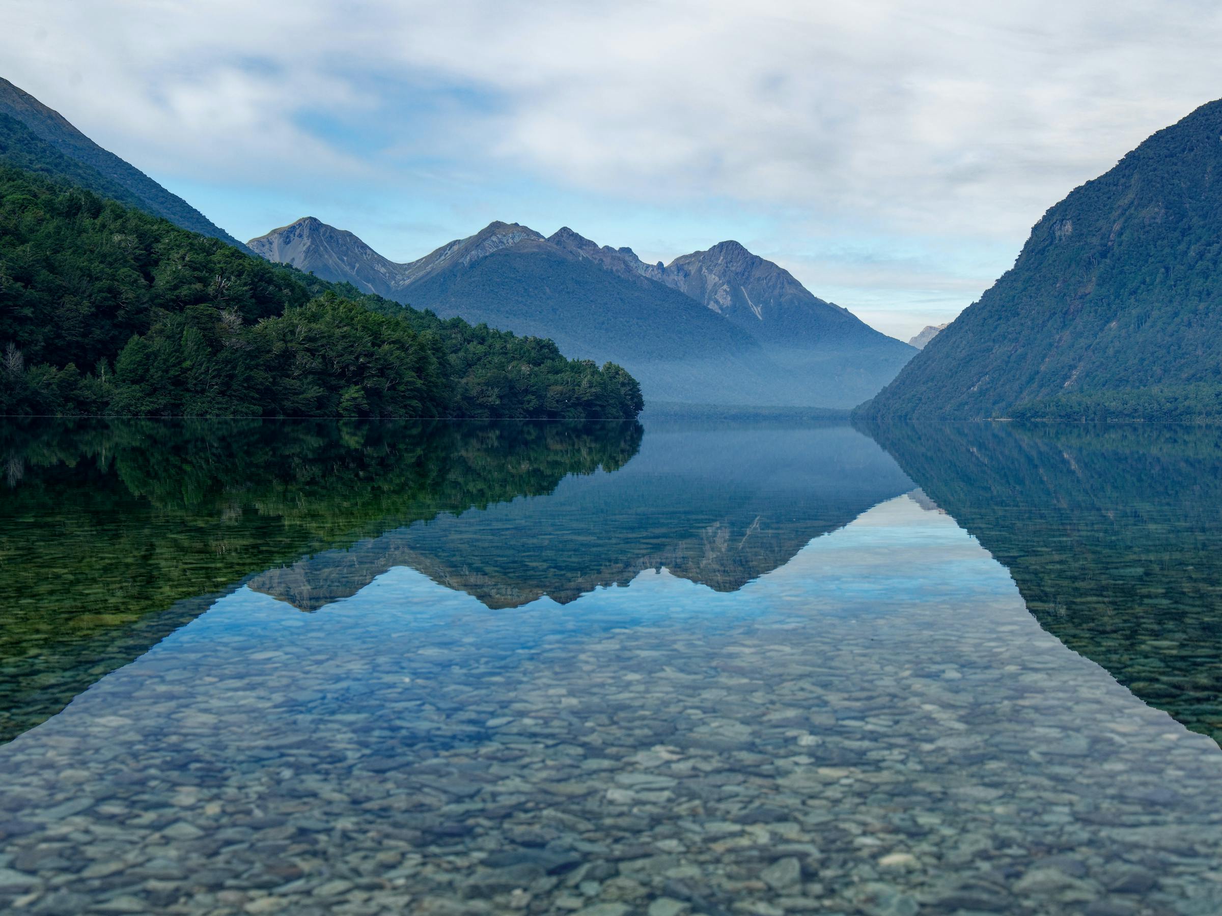 Still water at Gun Lake reflects forested slopes and misty peaks, the rocky shore visible through clear shallows.