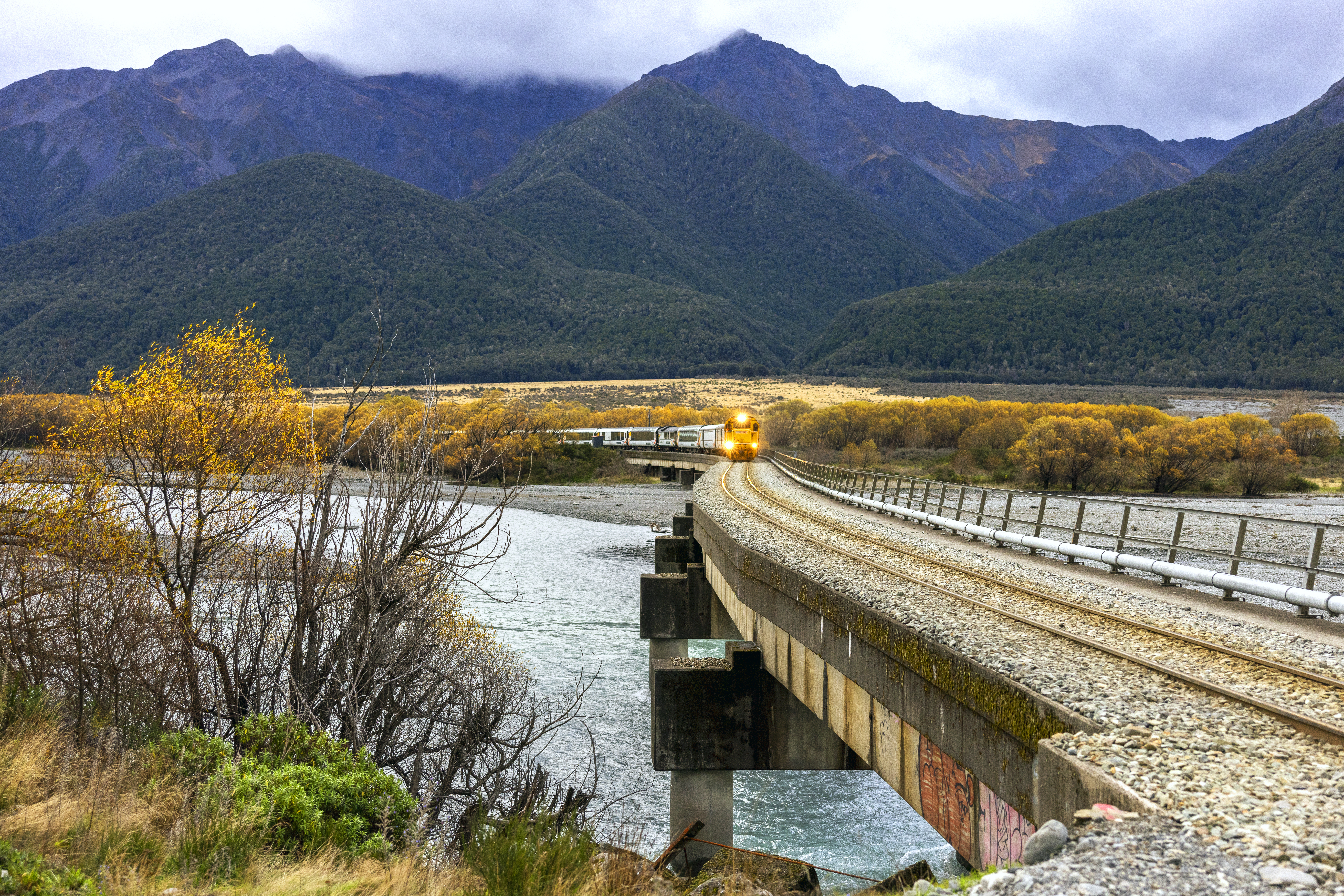 Milky turquoise river curves past a stone embankment and forested hillside, with low clouds over distant peaks.